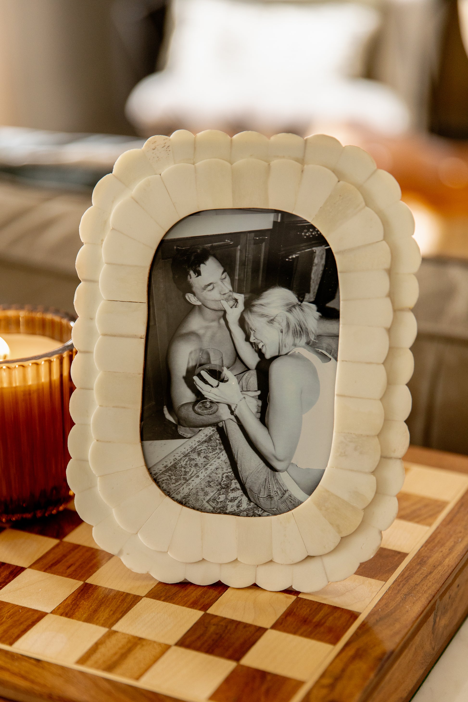 A black-and-white photo of a smiling couple is displayed in the Isla x Tate Roma 4X6 Oblong Picture Frame Ivory, featuring a scallop pattern and placed on a checkered wooden surface beside a textured glass candle holder.