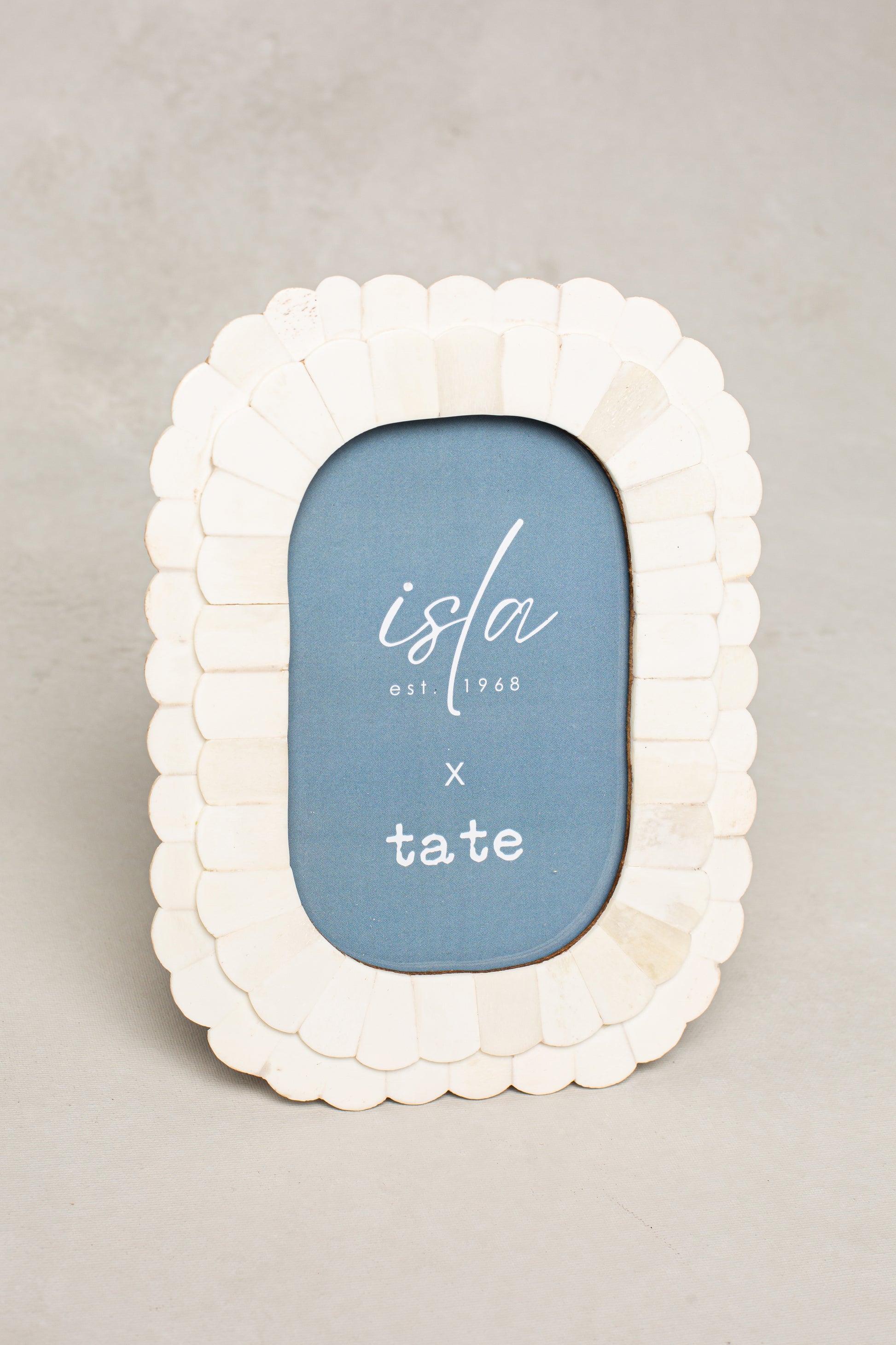 The Roma 4X6 Oblong Picture Frame Ivory by Isla x Tate features a white scallop pattern and blue insert with "isla est. 1968 x tate," shown on a light grey background.