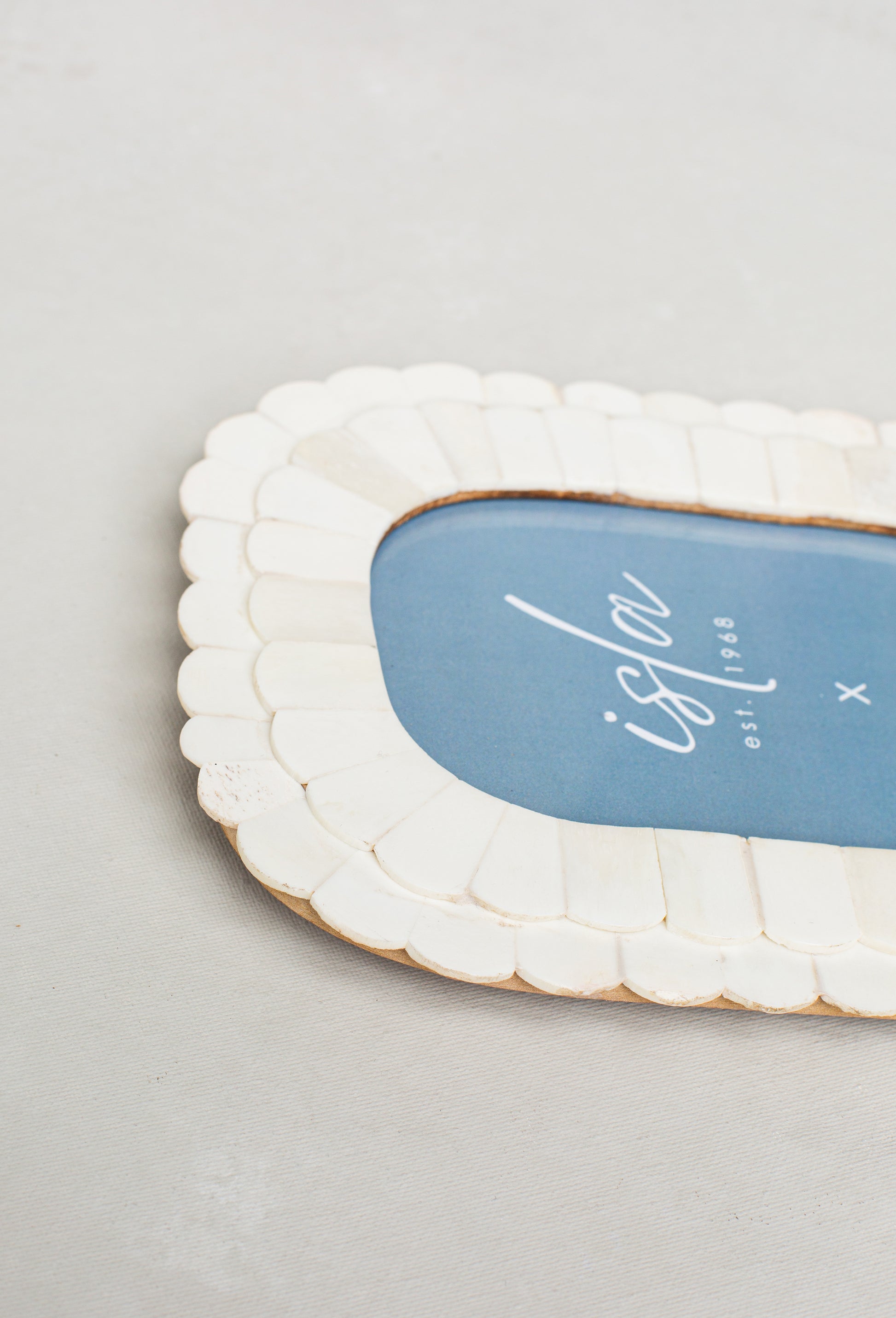 A close-up of the Roma 4X6 Oblong Picture Frame in Ivory by Isla x Tate, featuring a scalloped design and a blue center with "isla" and "est. 1988" in white script, displayed on a light-colored surface.