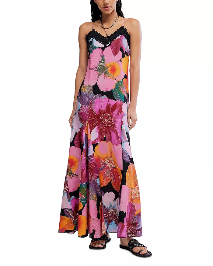 A person wears the Free People Bloomspell Maxi Dress in Pink Multi, featuring a bold floral print, spaghetti straps, black lace trim, and black sandals. Made from 100% viscose, shown against a plain white background.