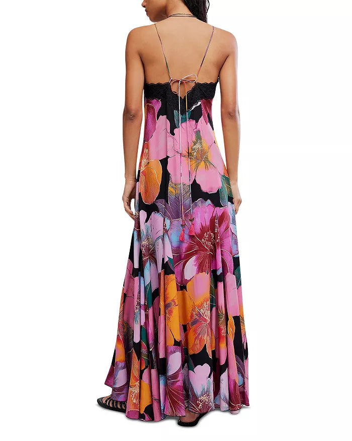 A woman is seen from behind wearing the Free People Bloomspell Maxi Dress, a long 100% viscose dress with spaghetti straps, a low open back with tie, and a vibrant floral print in pink, purple, orange, and yellow.