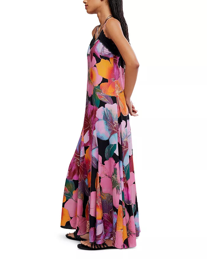 A woman stands sideways, hands clasped behind her back, wearing the Free People Bloomspell Maxi Dress—a colorful, sleeveless floral dress with spaghetti straps and a deep V-neck—crafted from 100% viscose for a breezy, elegant look.