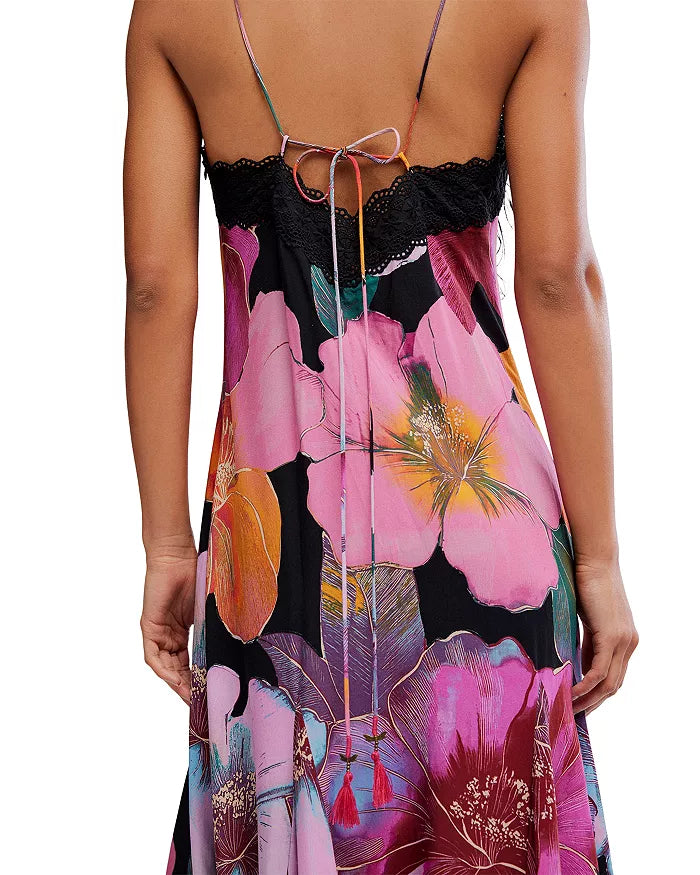 A woman is seen from behind wearing the Free People Bloomspell Maxi Dress—a sleeveless, open-back design with spaghetti straps tied in a bow and a vibrant pink, orange, purple, and black floral print.