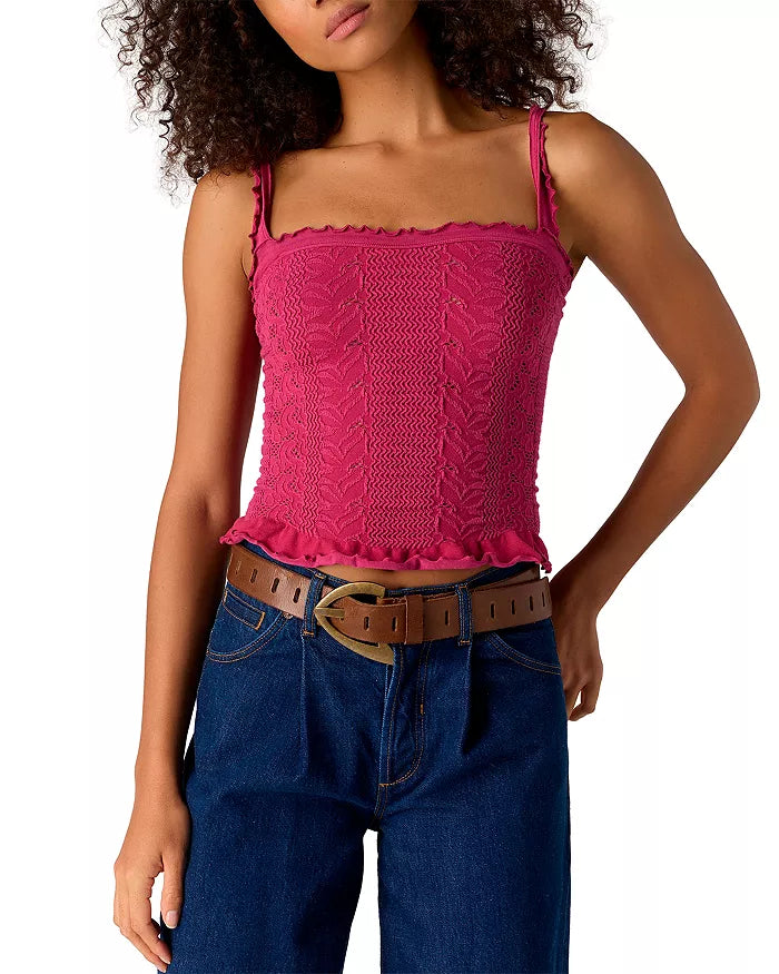 A woman wears the Free People So Soft Femme Square Top, a sleeveless magenta piece with ruffled edges, paired with blue jeans and a brown belt. She stands with one hand on her hip, her face partially out of frame.