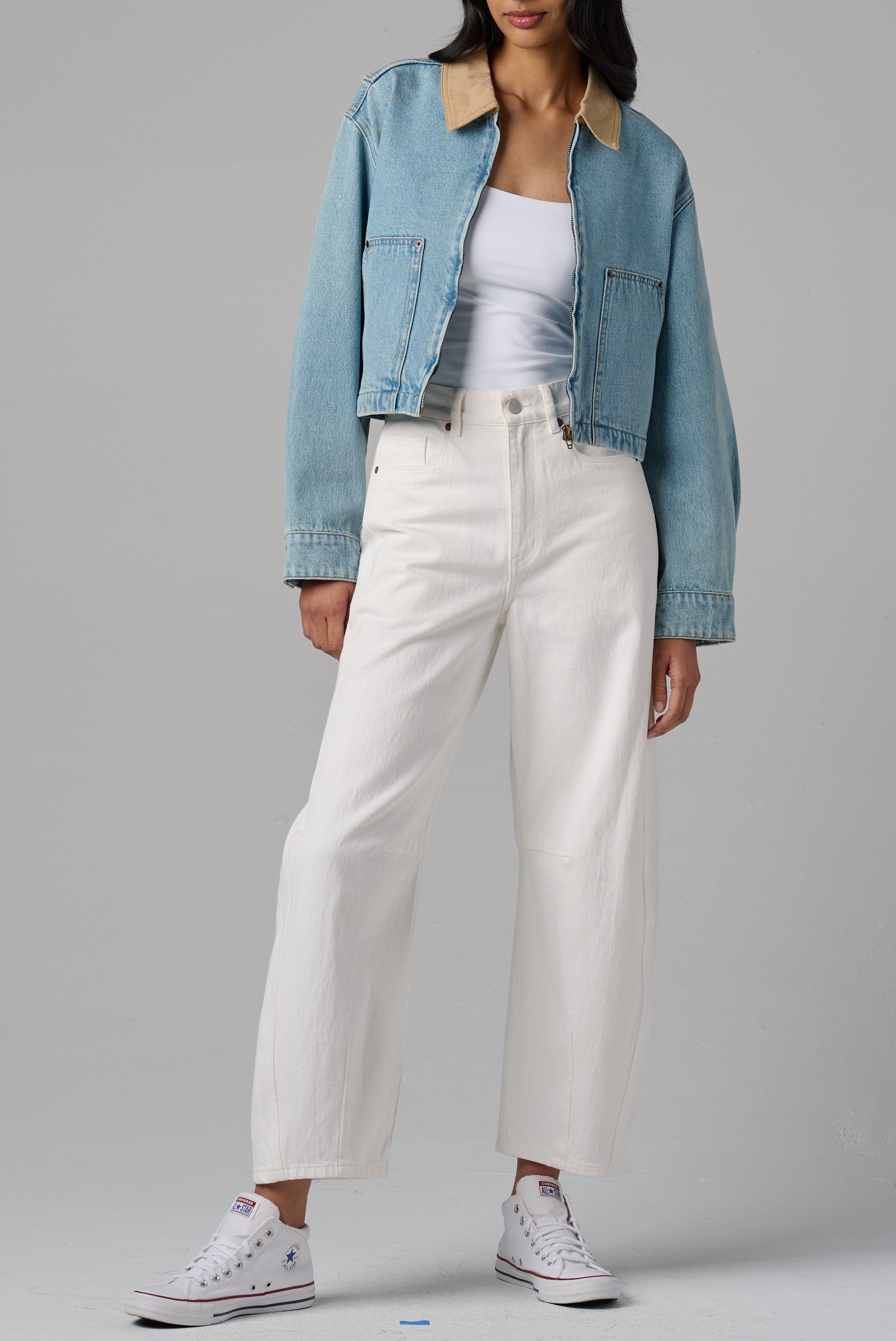A person stands against a gray background wearing Blank's Coffee Run Denim Barn Jacket in light blue with a beige collar and zip-front, paired with a white tank top, high-waisted white pants, and white Converse sneakers.