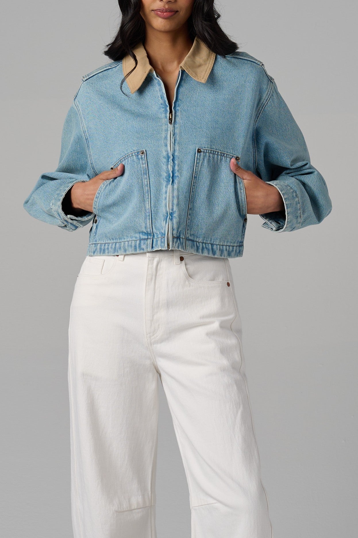 A woman stands against a gray background wearing the Coffee Run Denim Barn Jacket by Blank, featuring a light blue cropped fit, tan collar, zip-front closure, and pockets. She pairs it with white high-waisted pants; her head is partially cropped out.