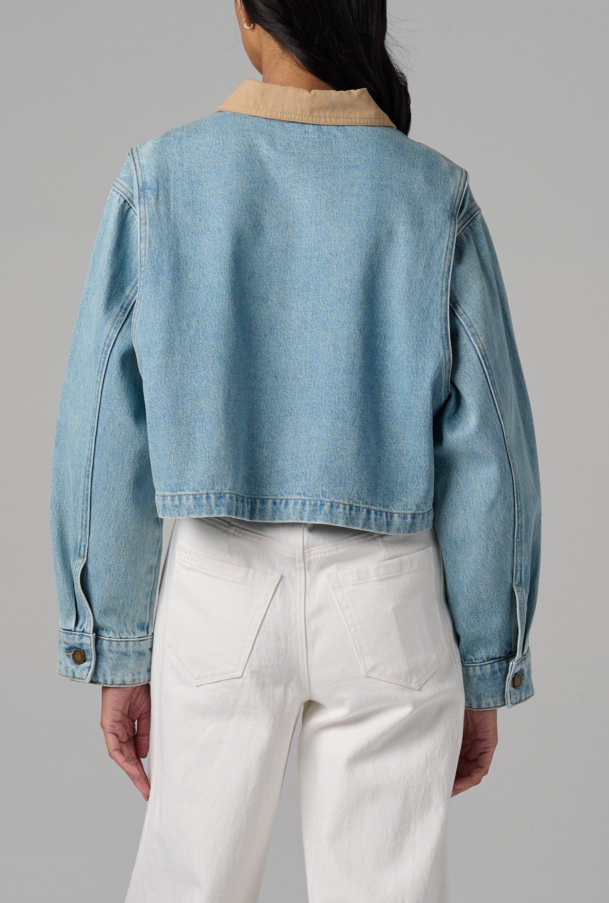 A person is seen from behind wearing Blank's Coffee Run Denim Barn Jacket, a light blue 100% cotton cropped style with a tan collar and buttoned cuffs, paired with high-waisted white pants against a plain gray background.