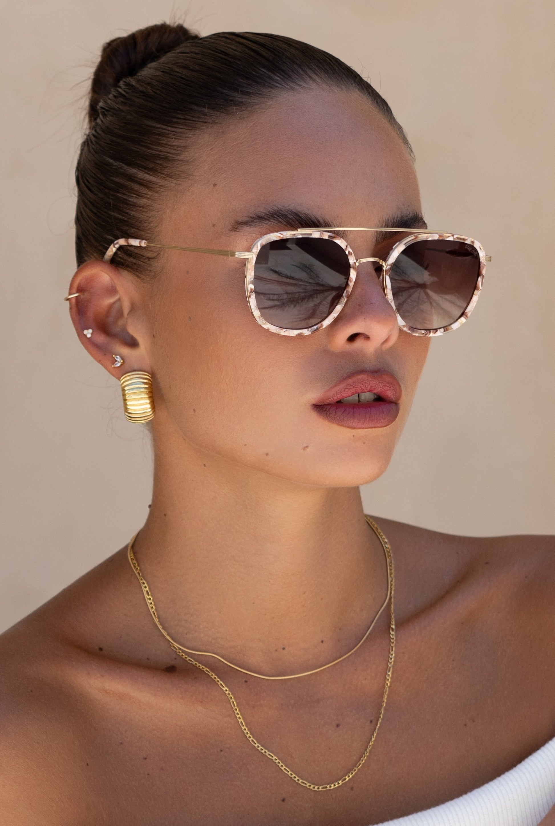 A woman in a white top and sleek bun poses against a beige background, wearing Freyrs Weston Sunnies in Pink Pearl/Brown with gold hoop earrings and layered gold necklaces.