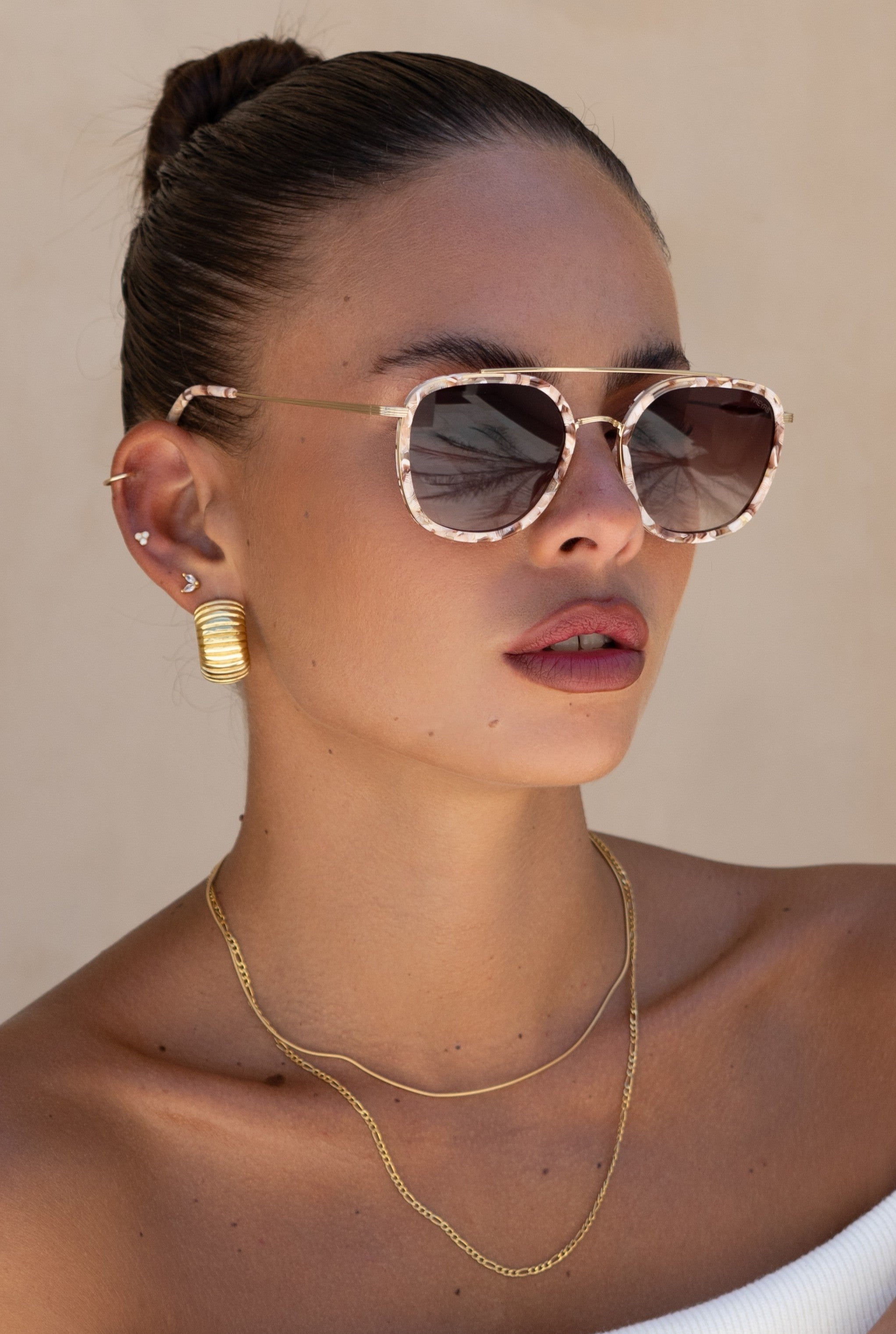 A woman in a white top and sleek bun poses against a beige background, wearing Freyrs Weston Sunnies in Pink Pearl/Brown with gold hoop earrings and layered gold necklaces.