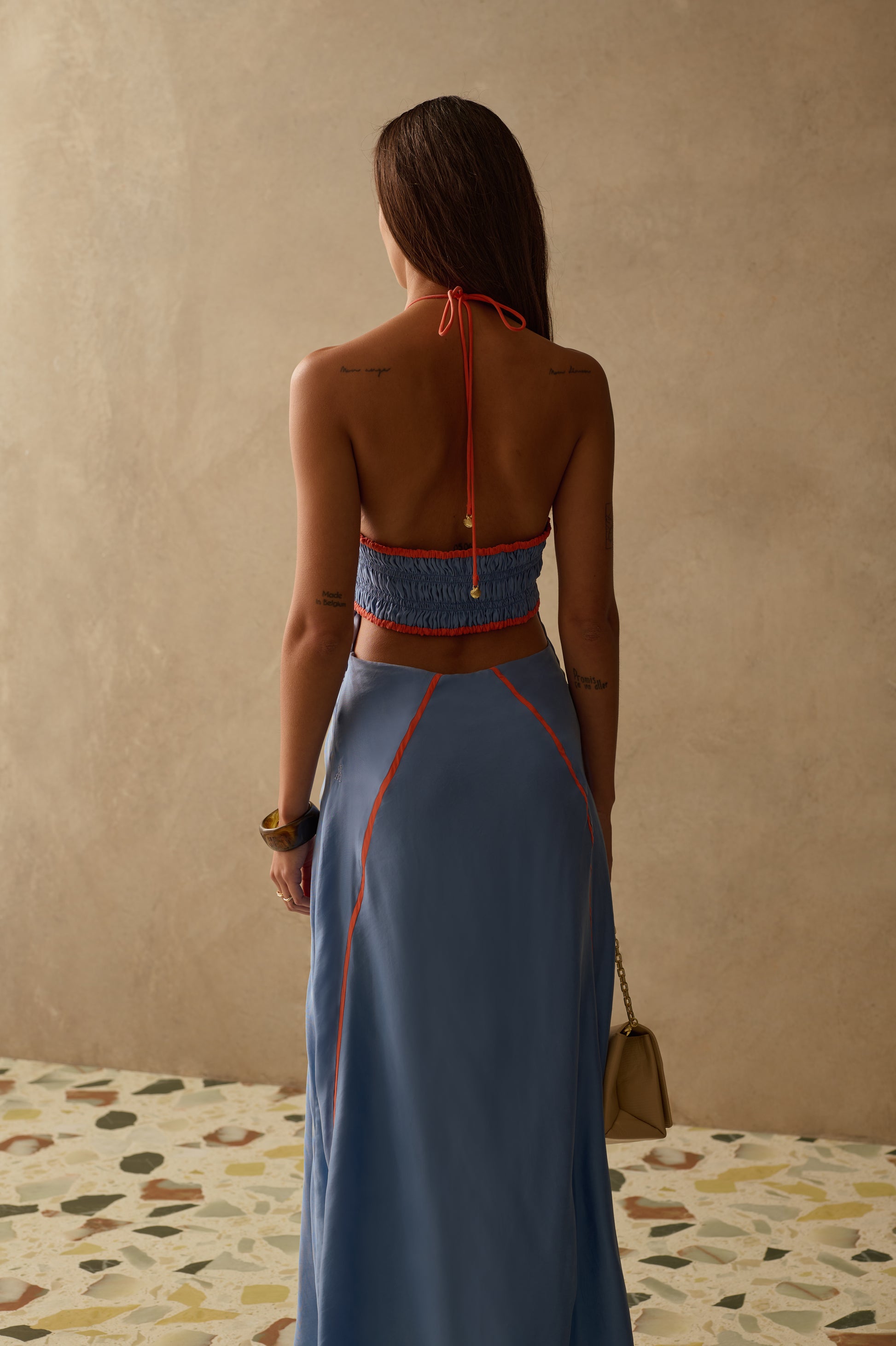 A woman with long brown hair stands facing away, wearing Sancia’s The Sloane Dress—a blue halter dress with a ruched neckline and red stitching. She holds a small beige handbag, on a terrazzo floor against a neutral beige wall.
