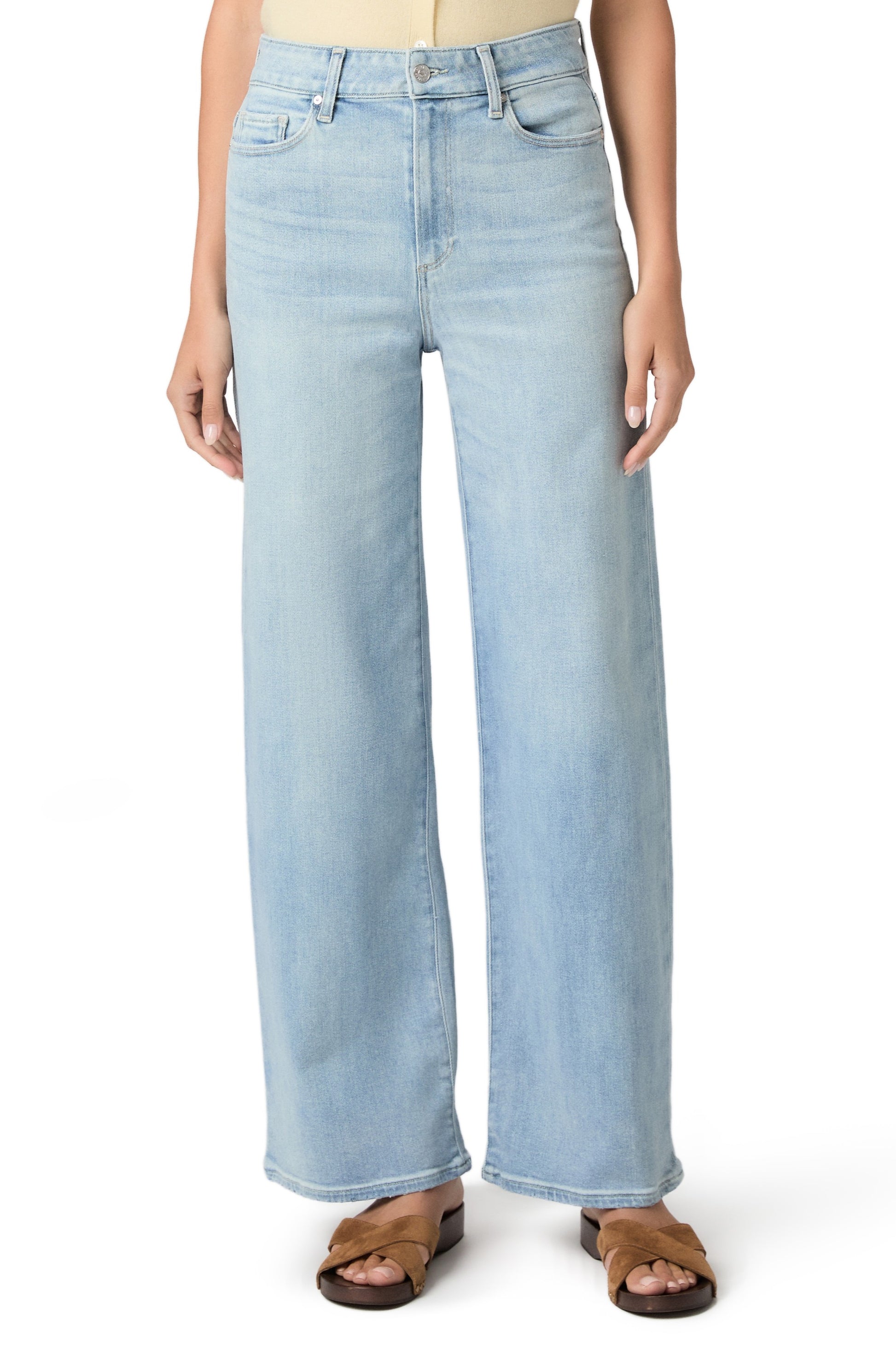 A person wears Paige's Anessa In Coliseum high-rise wide leg jeans in light denim with brown sandals, standing with arms relaxed at their sides; only the upper body is partially visible.