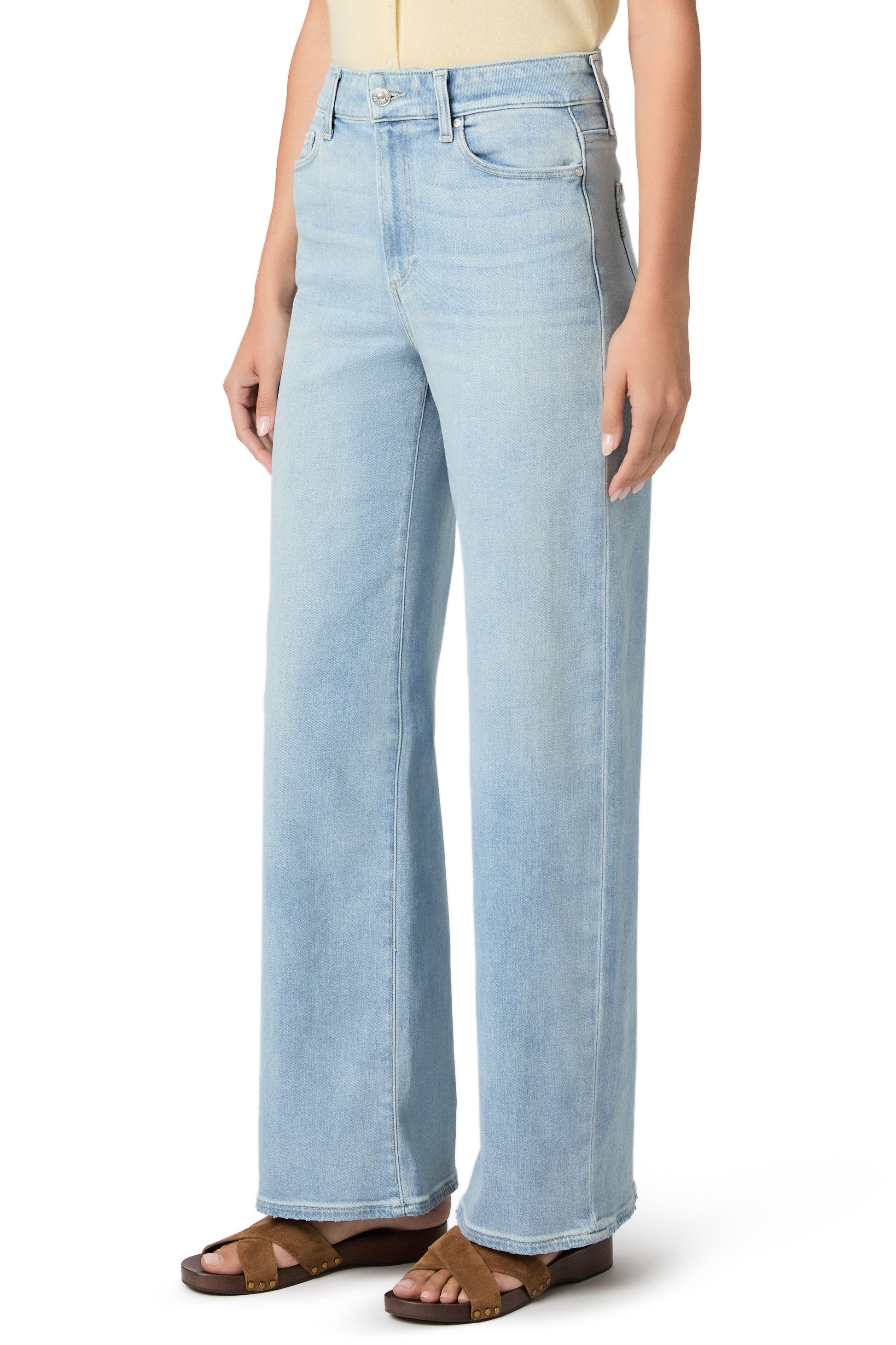 A person wearing Paige’s Anessa In Coliseum high-rise wide leg jeans in light blue, paired with a tucked-in light yellow top and brown sandals, stands against a plain white background.