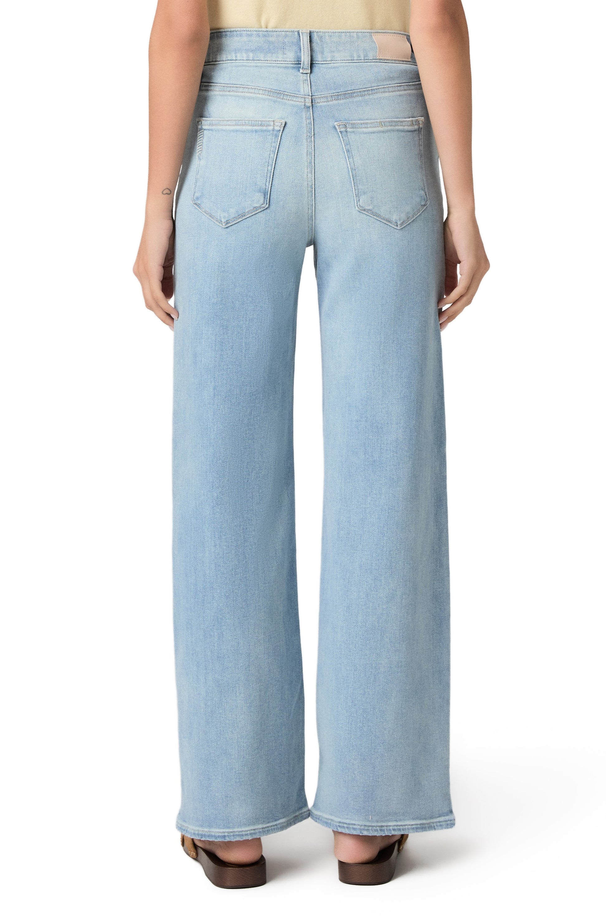 A person is shown from the back against a white background, wearing Paige Anessa In Coliseum high-rise wide leg jeans in a light blue wash and brown sandals.