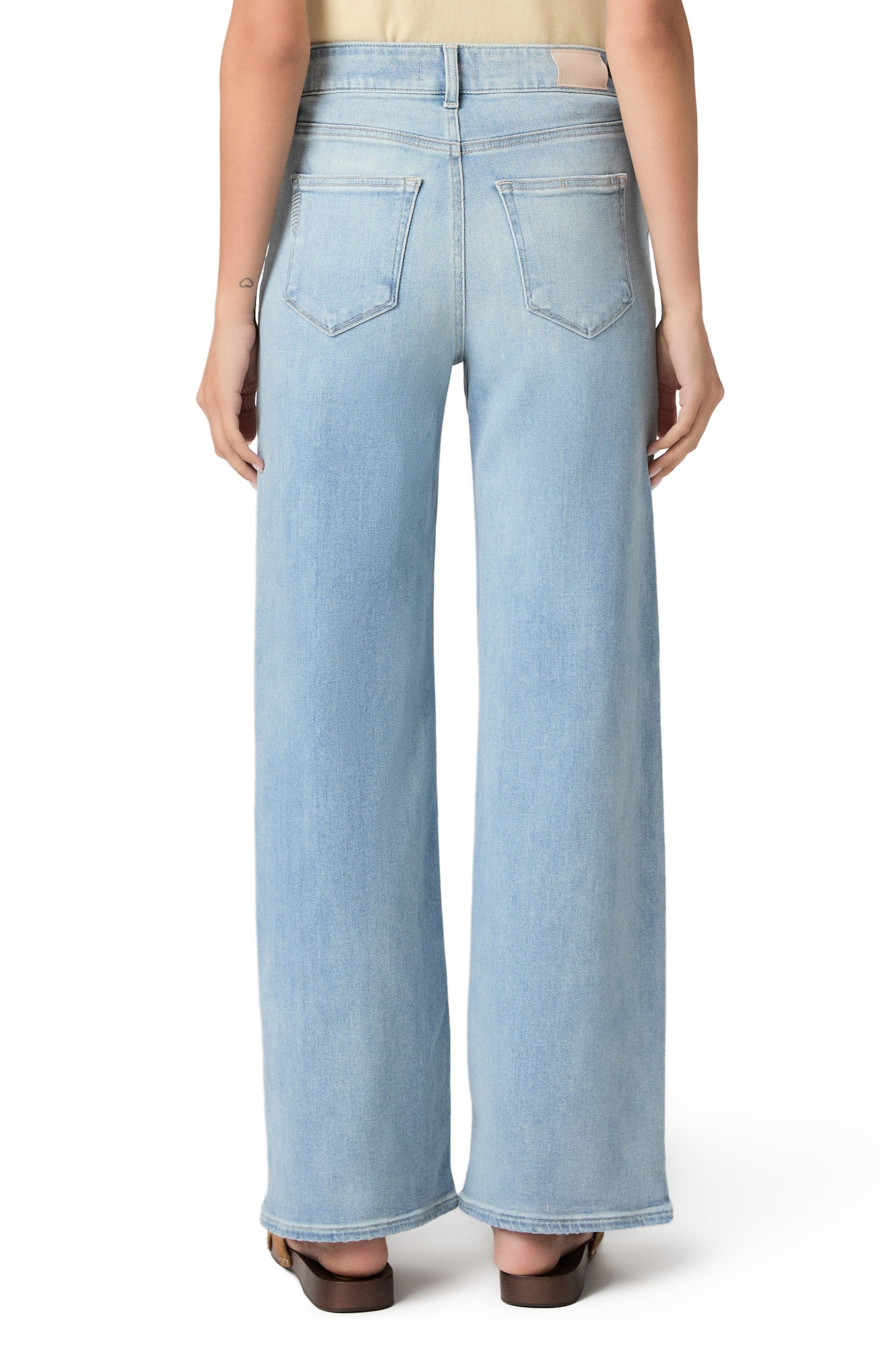 A person is shown from the back against a white background, wearing Paige Anessa In Coliseum high-rise wide leg jeans in a light blue wash and brown sandals.