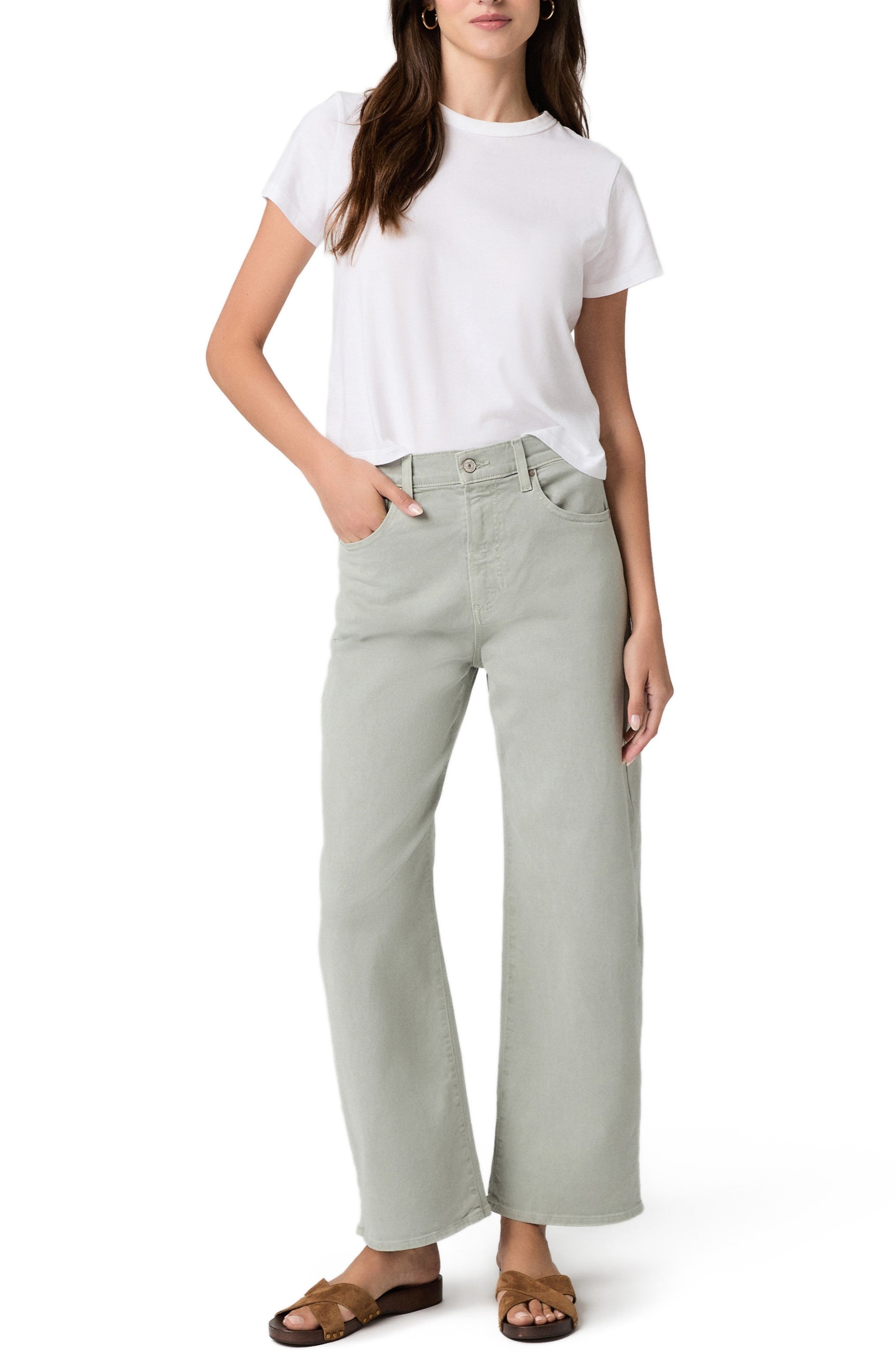 A woman stands with one hand in her pocket, wearing the Paige Ariella In Sea Glass high rise barrel leg pants, a white t-shirt, and brown sandals against a plain white background.
