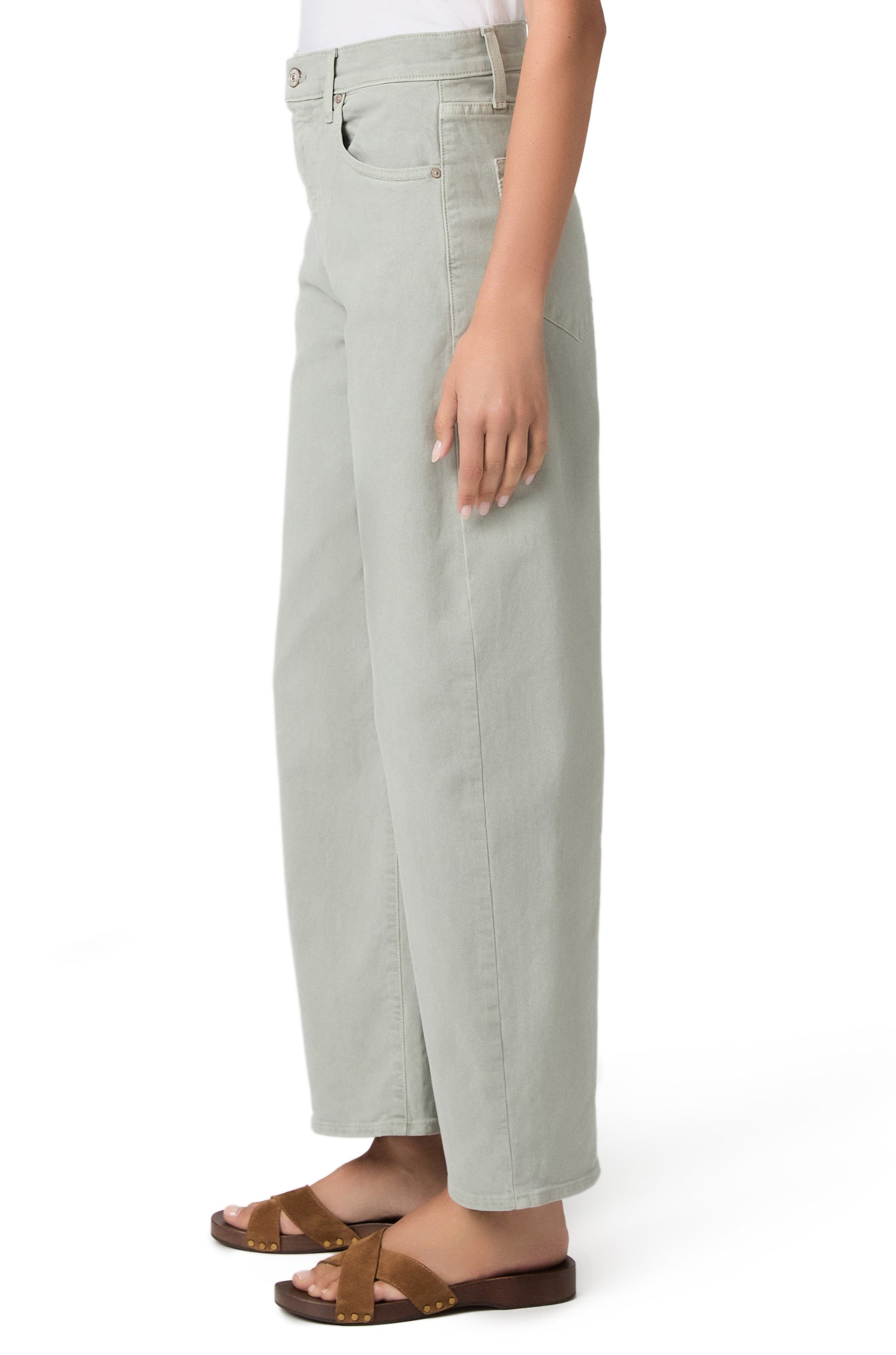 A person is shown from the waist down, standing sideways against a white background, wearing Paige’s Ariella In Sea Glass jeans and brown sandals.