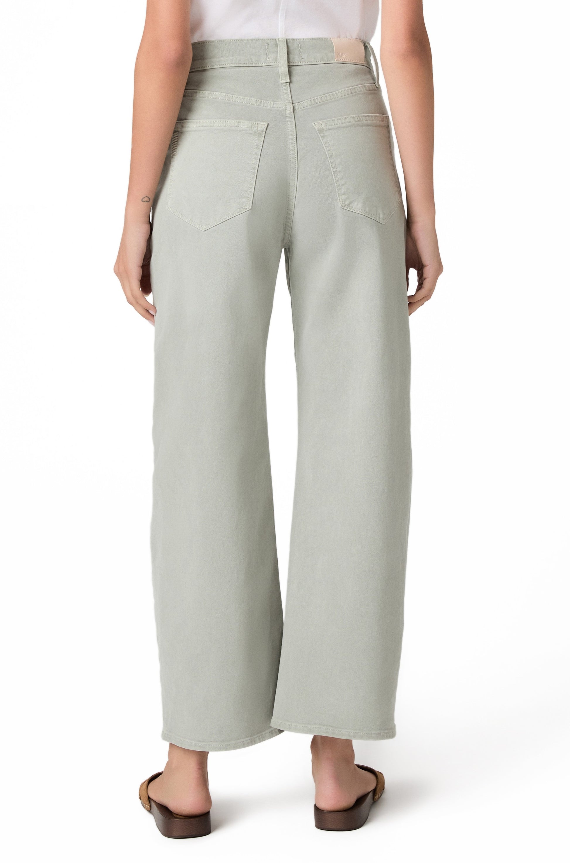 A person stands with their back to the camera, wearing Paige's Ariella In Sea Glass jeans—a light green barrel leg style—paired with a white tucked-in shirt and brown sandals. Only the lower half is visible against a white background.