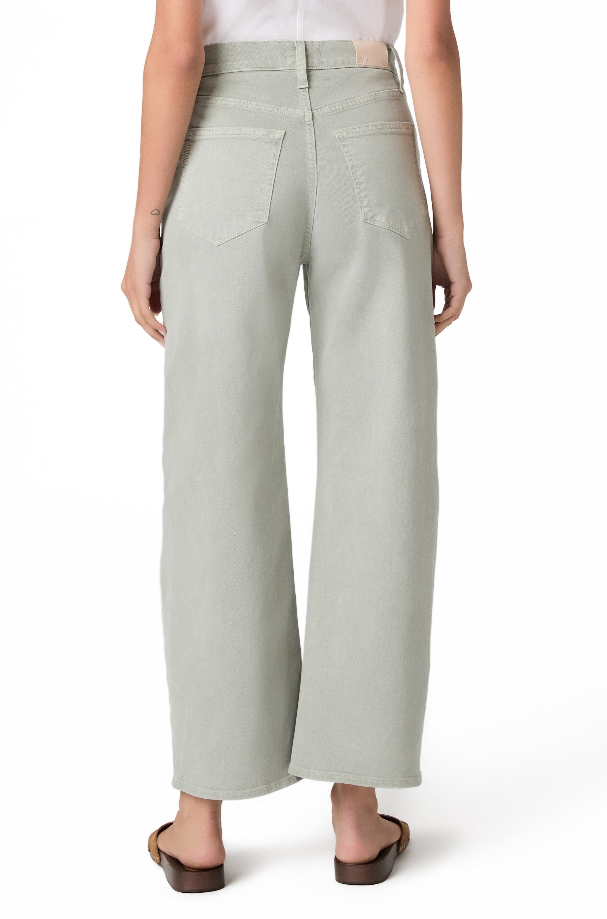 A person stands with their back to the camera, wearing Paige's Ariella In Sea Glass jeans—a light green barrel leg style—paired with a white tucked-in shirt and brown sandals. Only the lower half is visible against a white background.