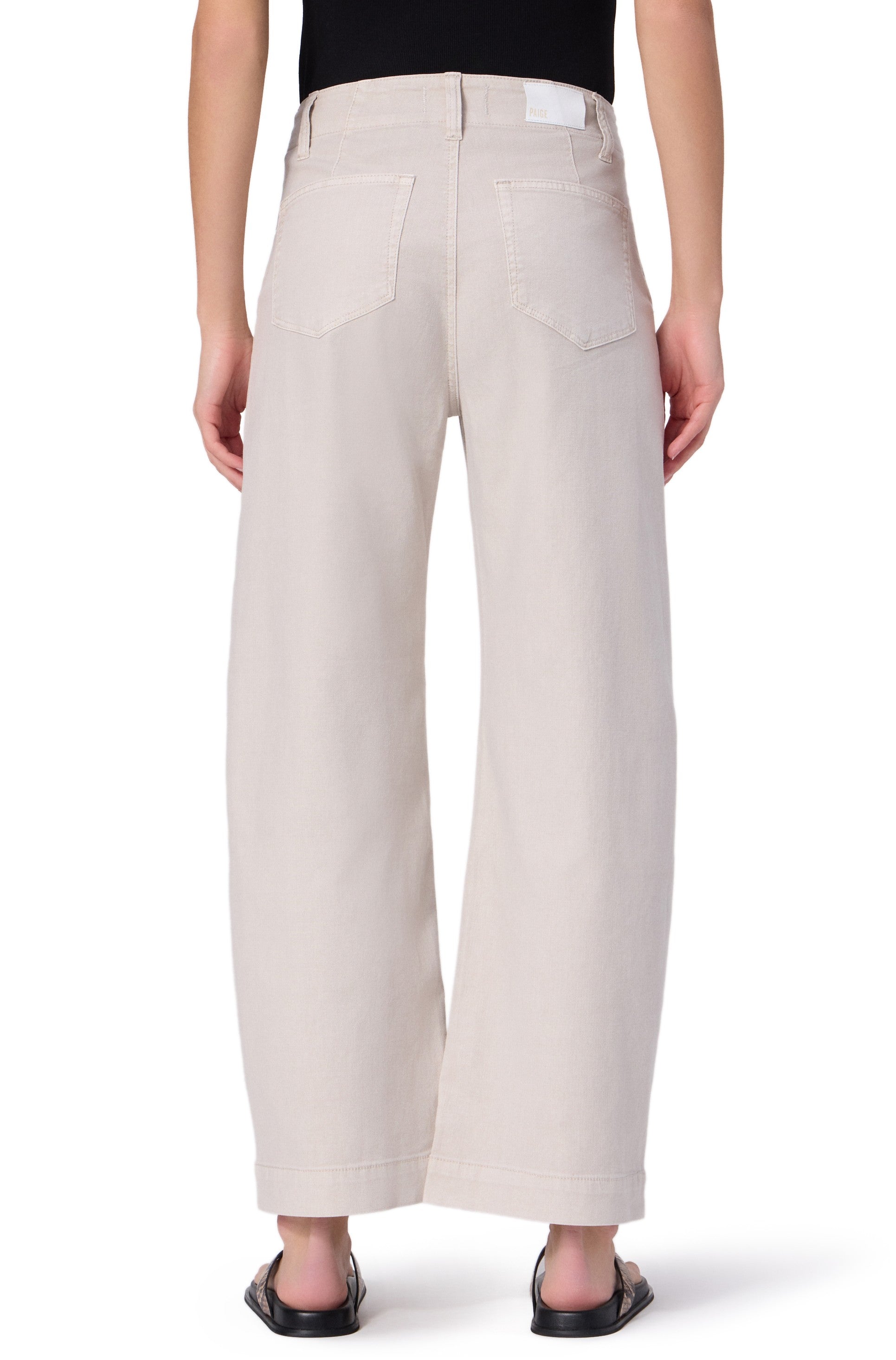 A person wearing Paige Arellia Ankle With Tie relaxed fit light beige wide-leg jeans and black sandals, shown from the back. The soft denim pants have two back pockets, a white waistband label, and are styled with a black top.