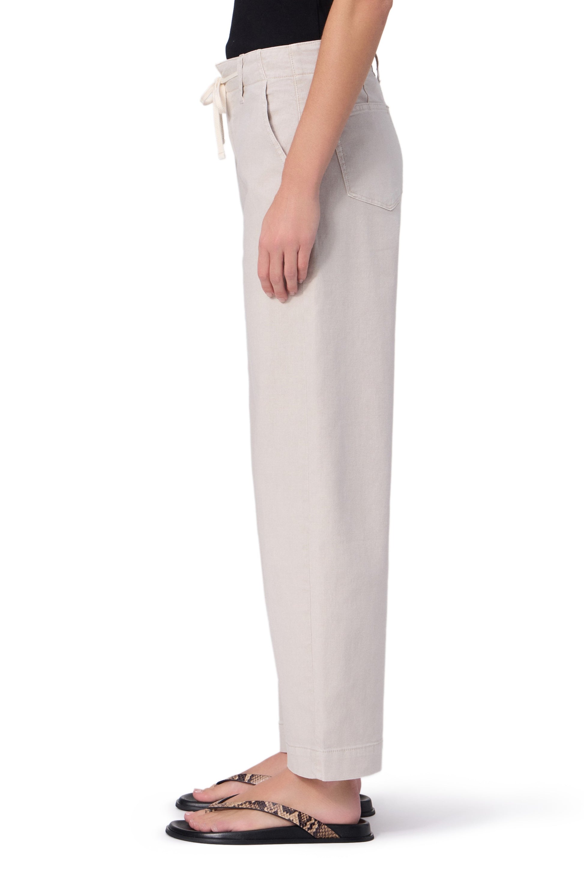 A person stands in profile against a white background, wearing Paige’s Arellia Ankle With Tie relaxed fit light beige soft denim pants, paired with a black top and snakeskin-patterned flip-flops.