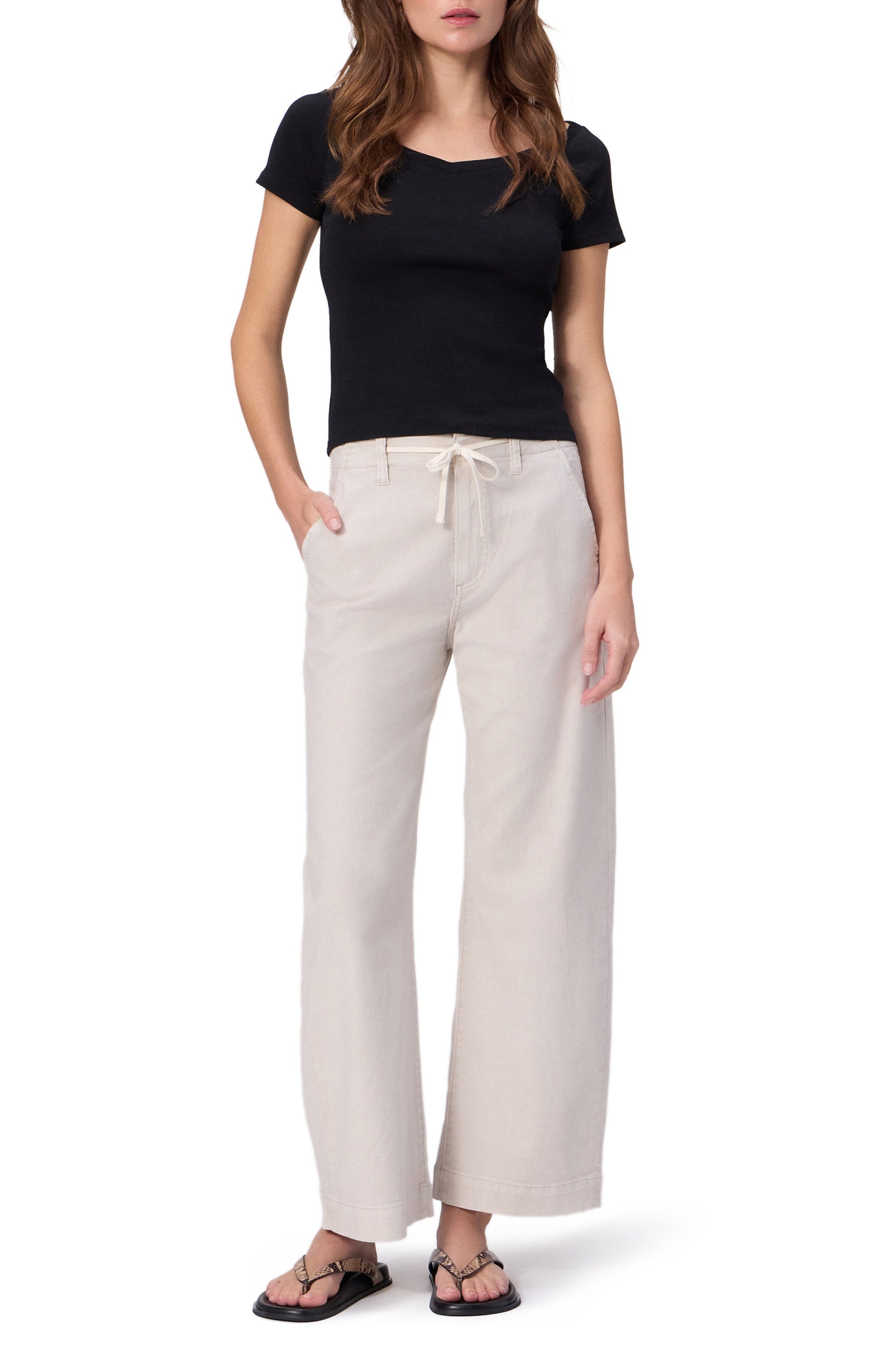 A person stands with hands in pockets, wearing a black short-sleeve top and Paige Arellia Ankle With Tie light beige relaxed fit soft denim pants, paired with black sandals. The photo is cropped above the knees.
