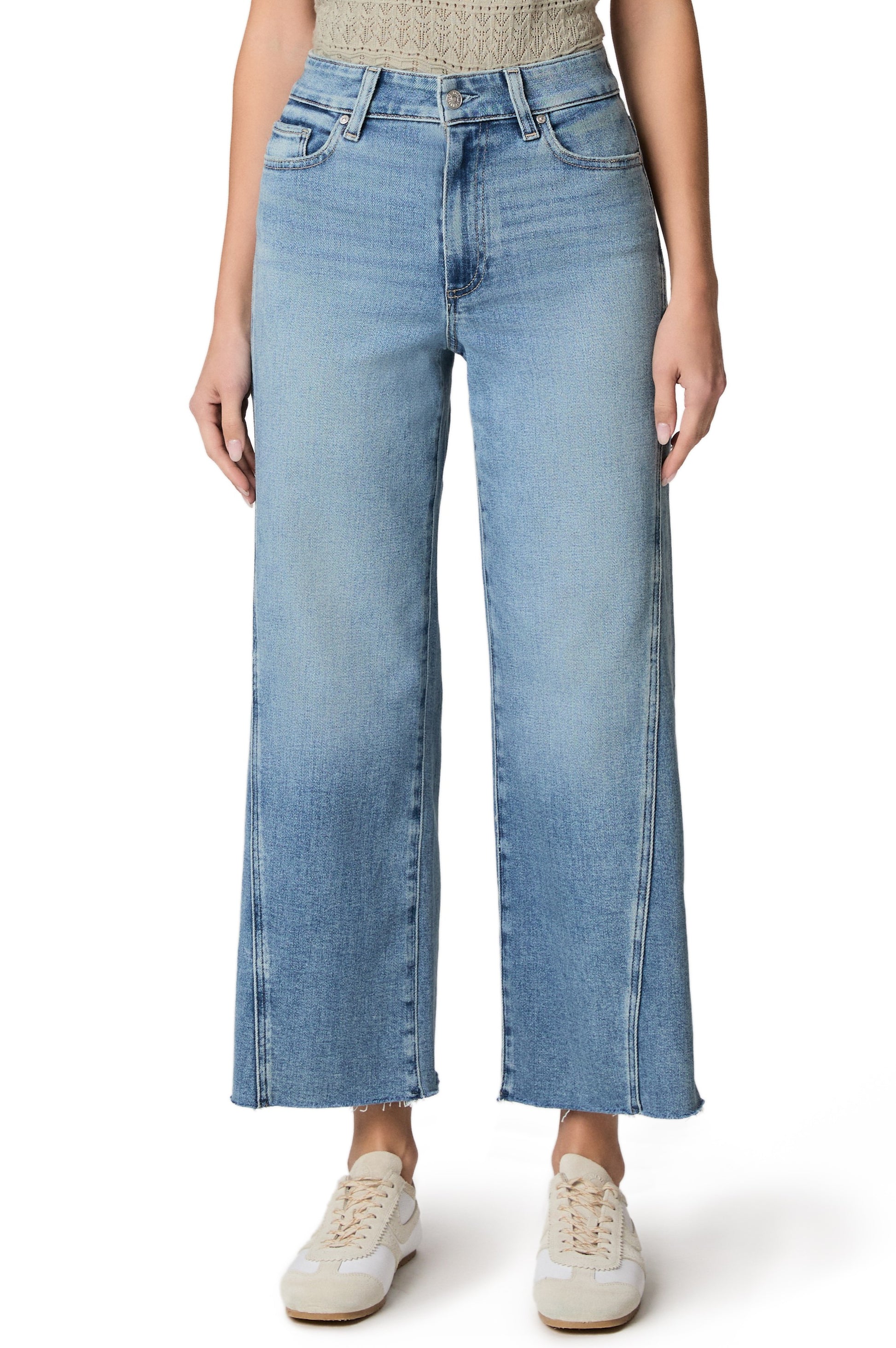 A person stands against a white background wearing Paige's Anessa with Twisted Outseam Panel + Raw Hem in light denim, styled with a sleeveless knit top and white sneakers.