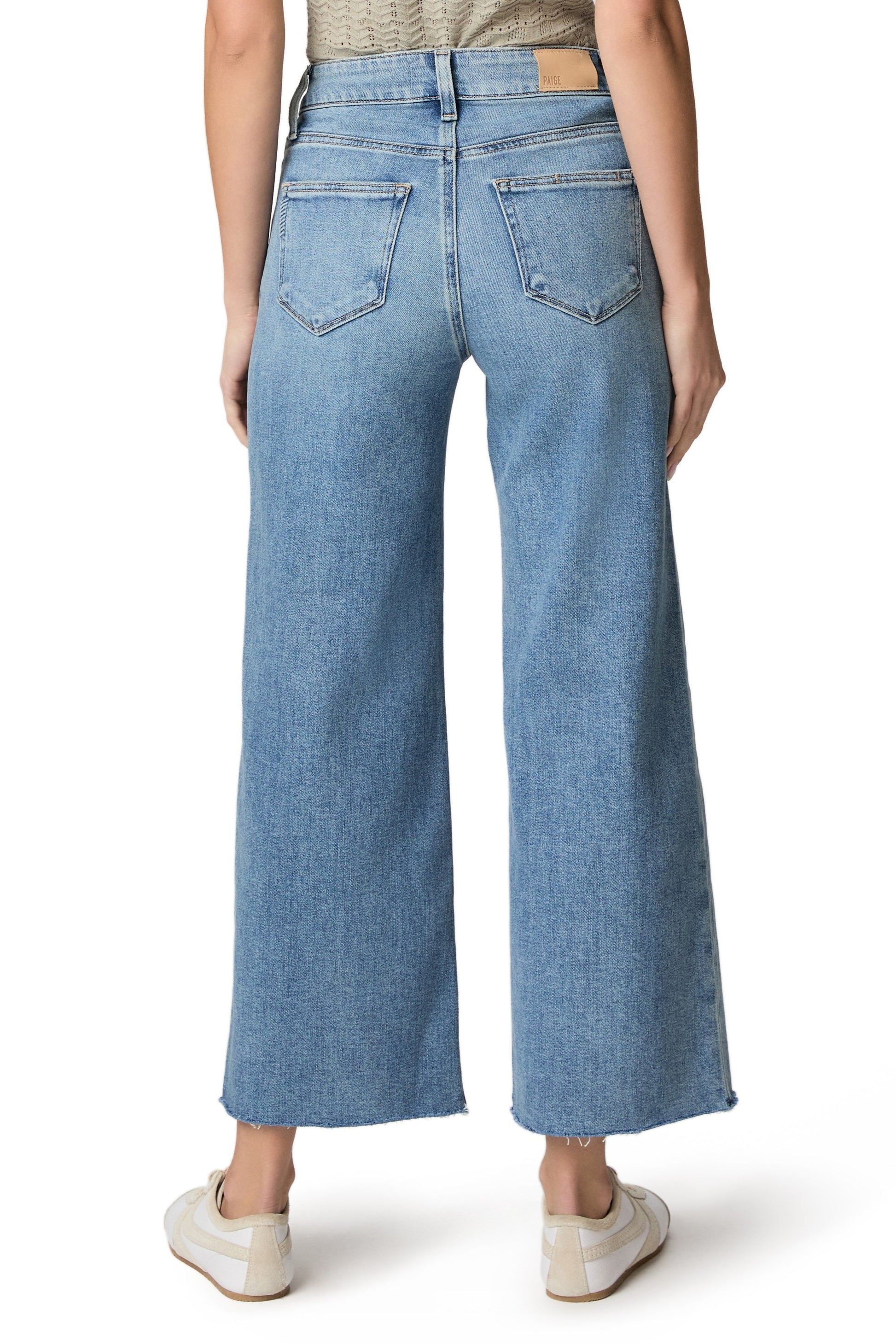 A person wearing the Paige Anessa jeans with a twisted outseam panel and raw hem in a relaxed fit, light blue wash, paired with white sneakers, is shown from the back against a white background.