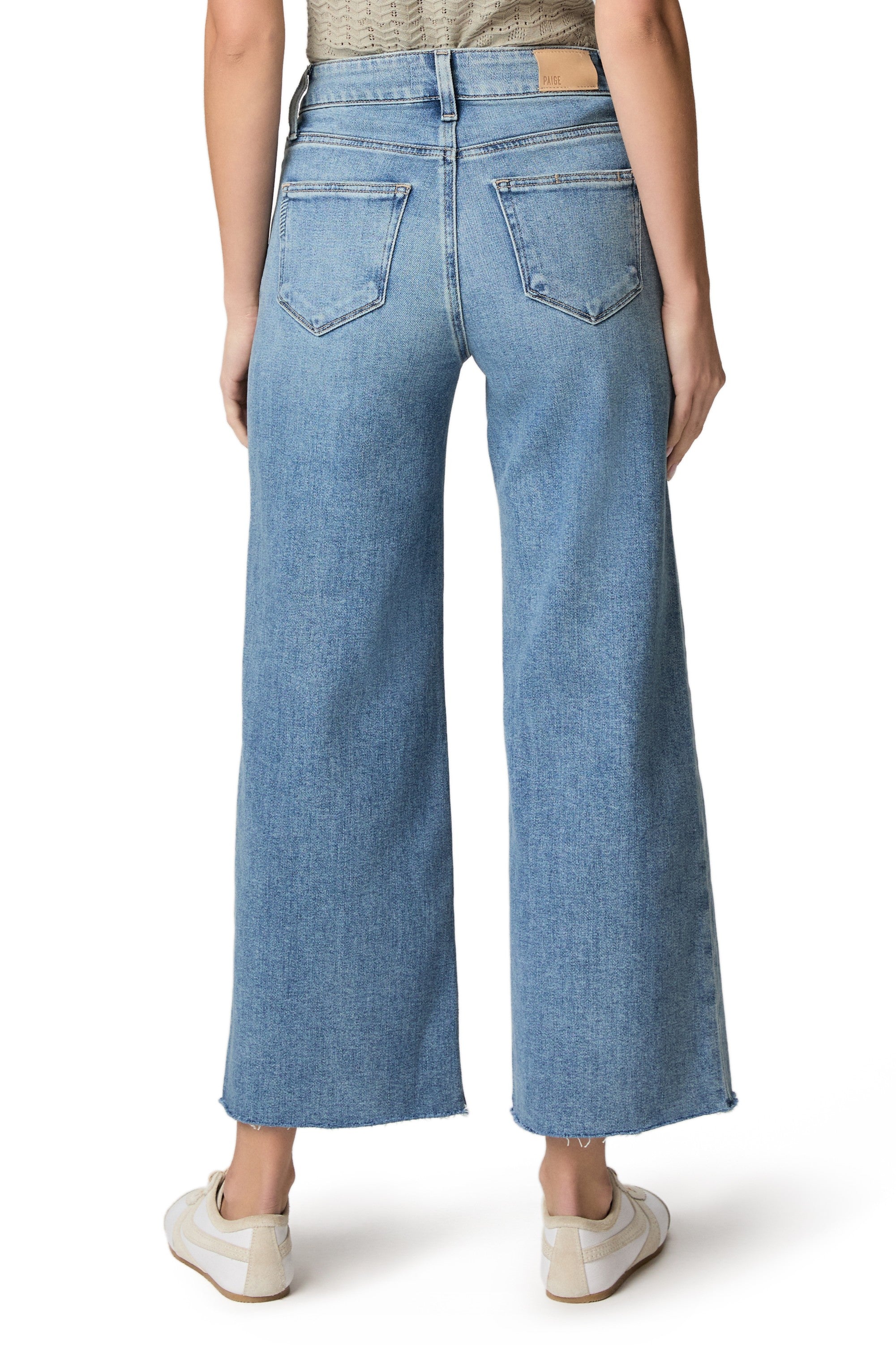 A person wearing the Paige Anessa jeans with a twisted outseam panel and raw hem in a relaxed fit, light blue wash, paired with white sneakers, is shown from the back against a white background.