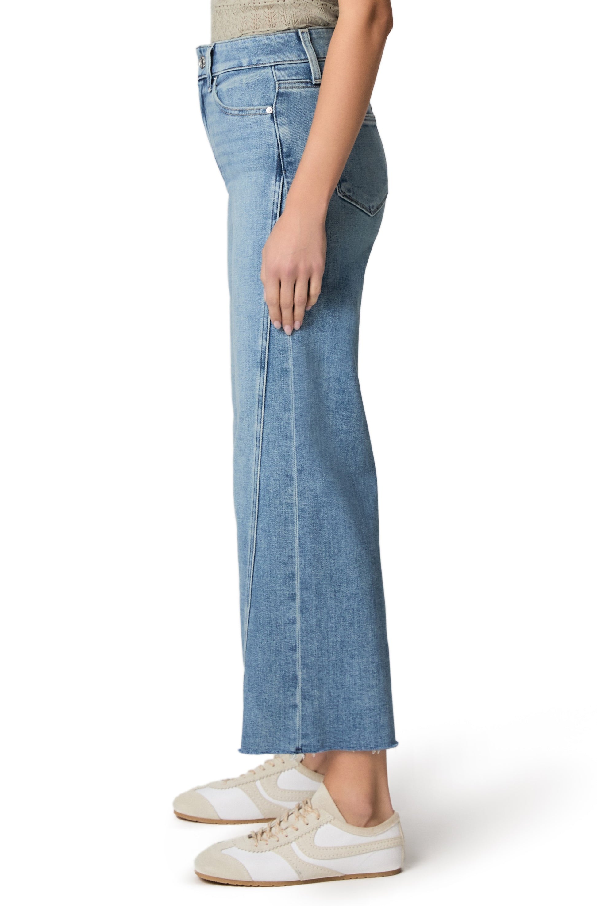 A person wearing Paige's Anessa with Twisted Outseam Panel + Raw Hem jeans, a beige top, and white sneakers stands sideways against a white background.