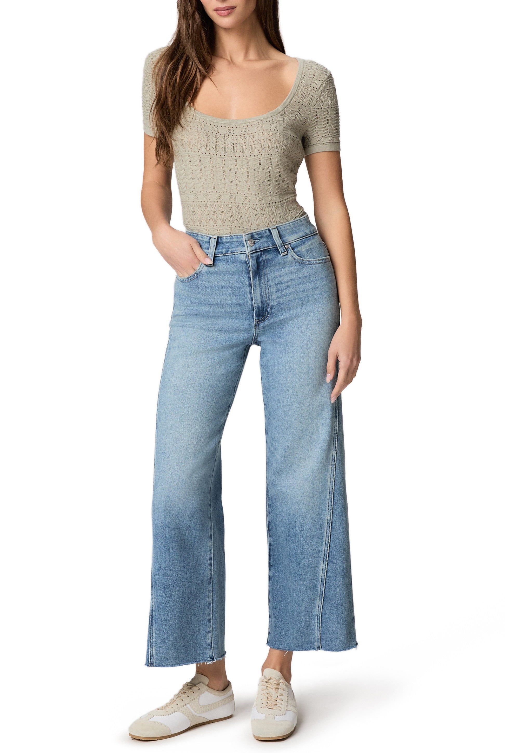 A woman in a short-sleeve beige knit top, Paige Anessa with Twisted Outseam Panel + Raw Hem jeans, and white sneakers stands with one hand in her pocket against a plain white background.