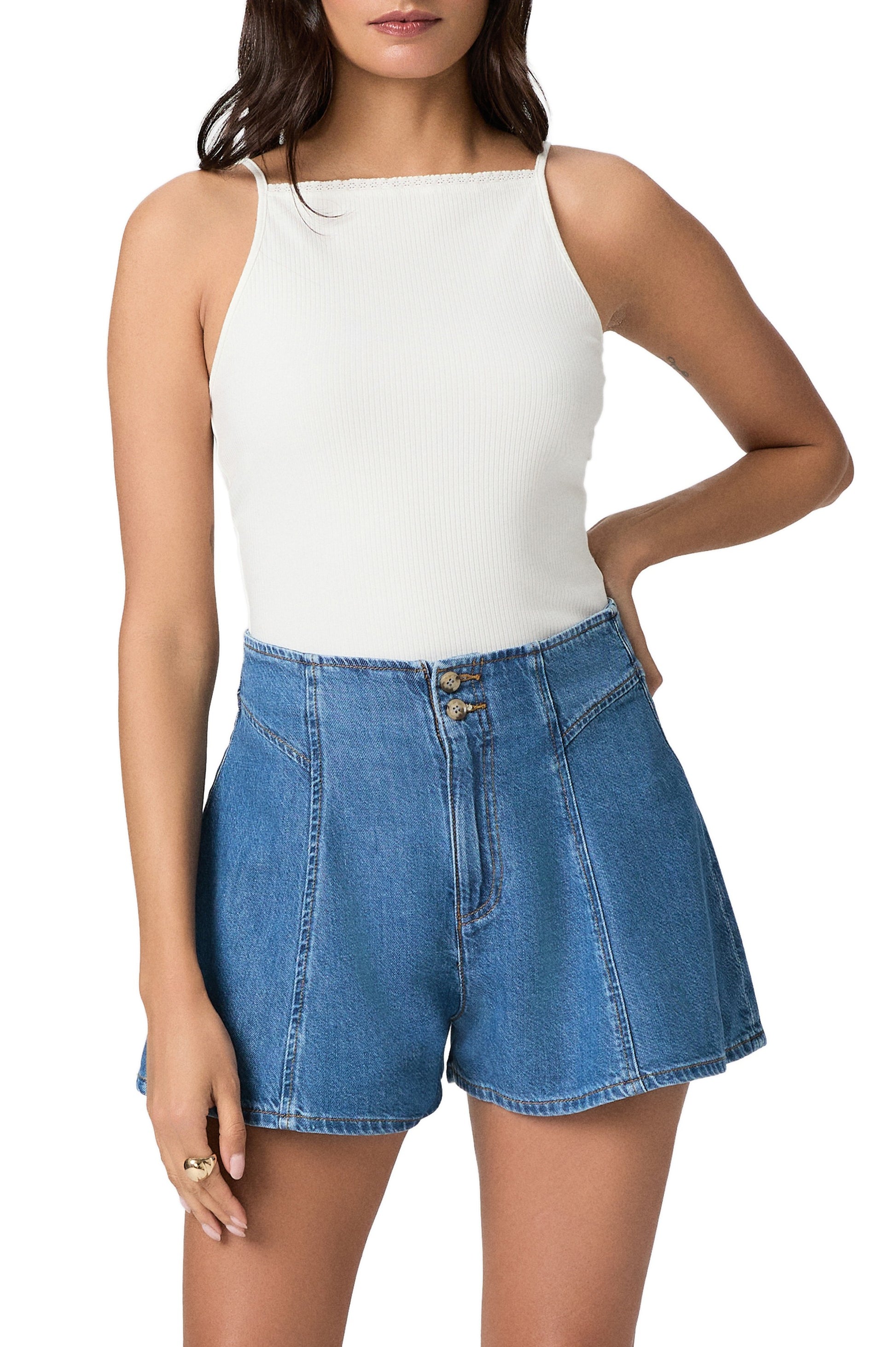A woman wears a sleeveless white ribbed top and Paige Joelle Short - Anchored, high-waisted lightweight denim shorts with front seams, standing with one hand on her hip against a white background.
