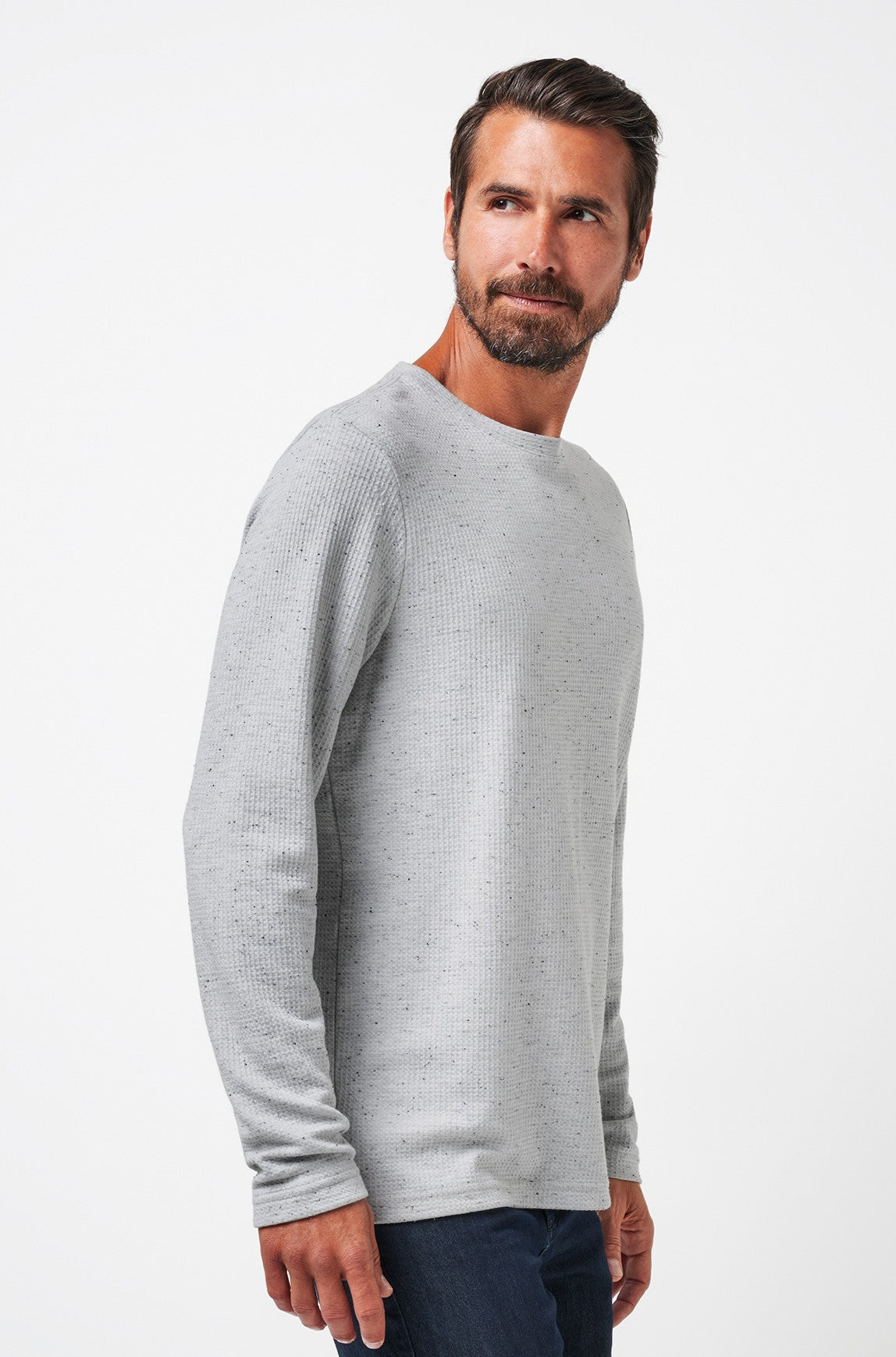 A man with short brown hair and a beard wears a light grey TravisMathew Cloud Waffle Crew and dark jeans, standing against a plain white background and looking slightly to the side.
