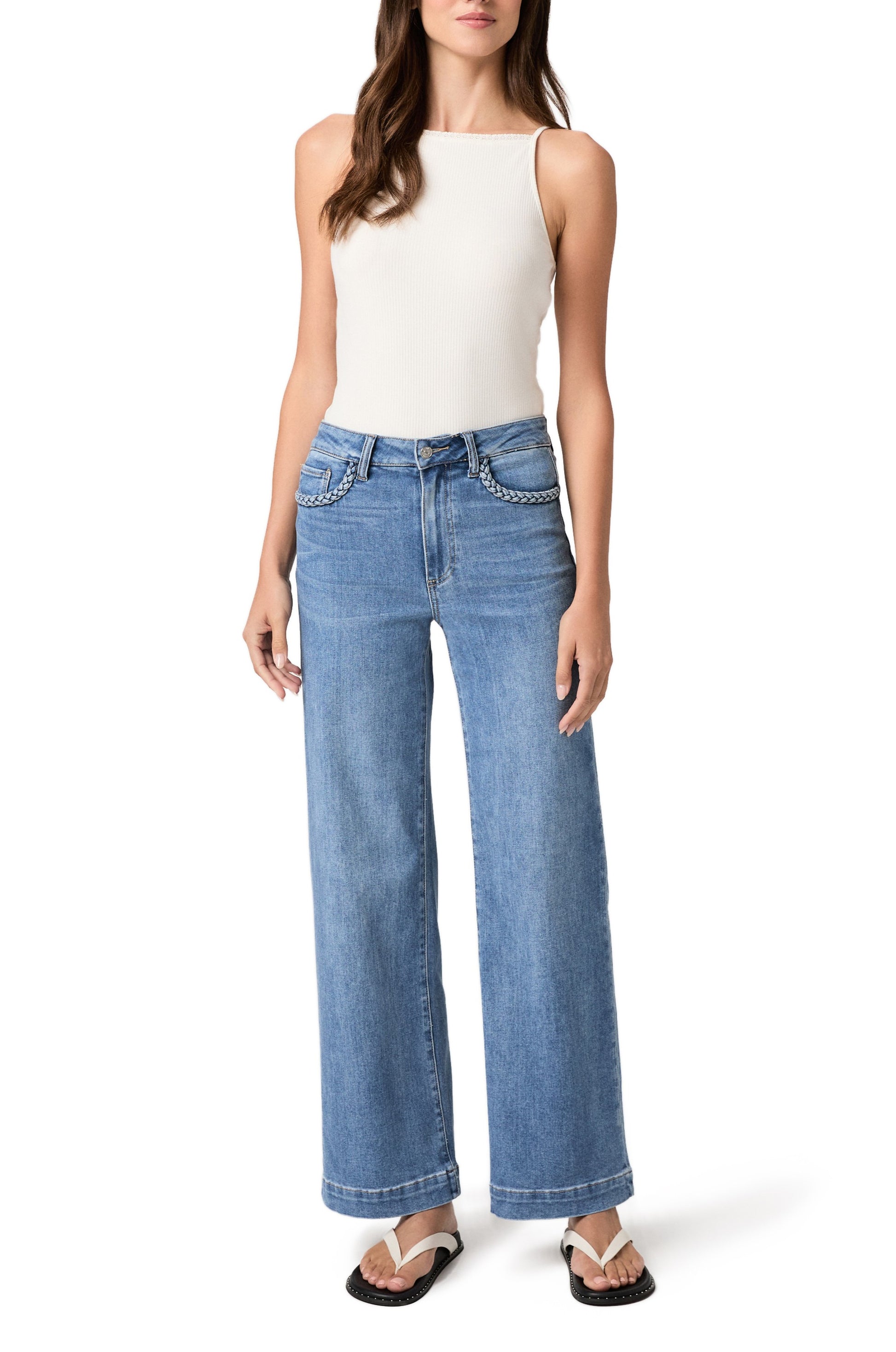 Woman wearing a white sleeveless top, Paige Anessa 31" denim high rise wide leg jeans with braided pocket edge in the Encino wash, and black sandals, standing against a white background.