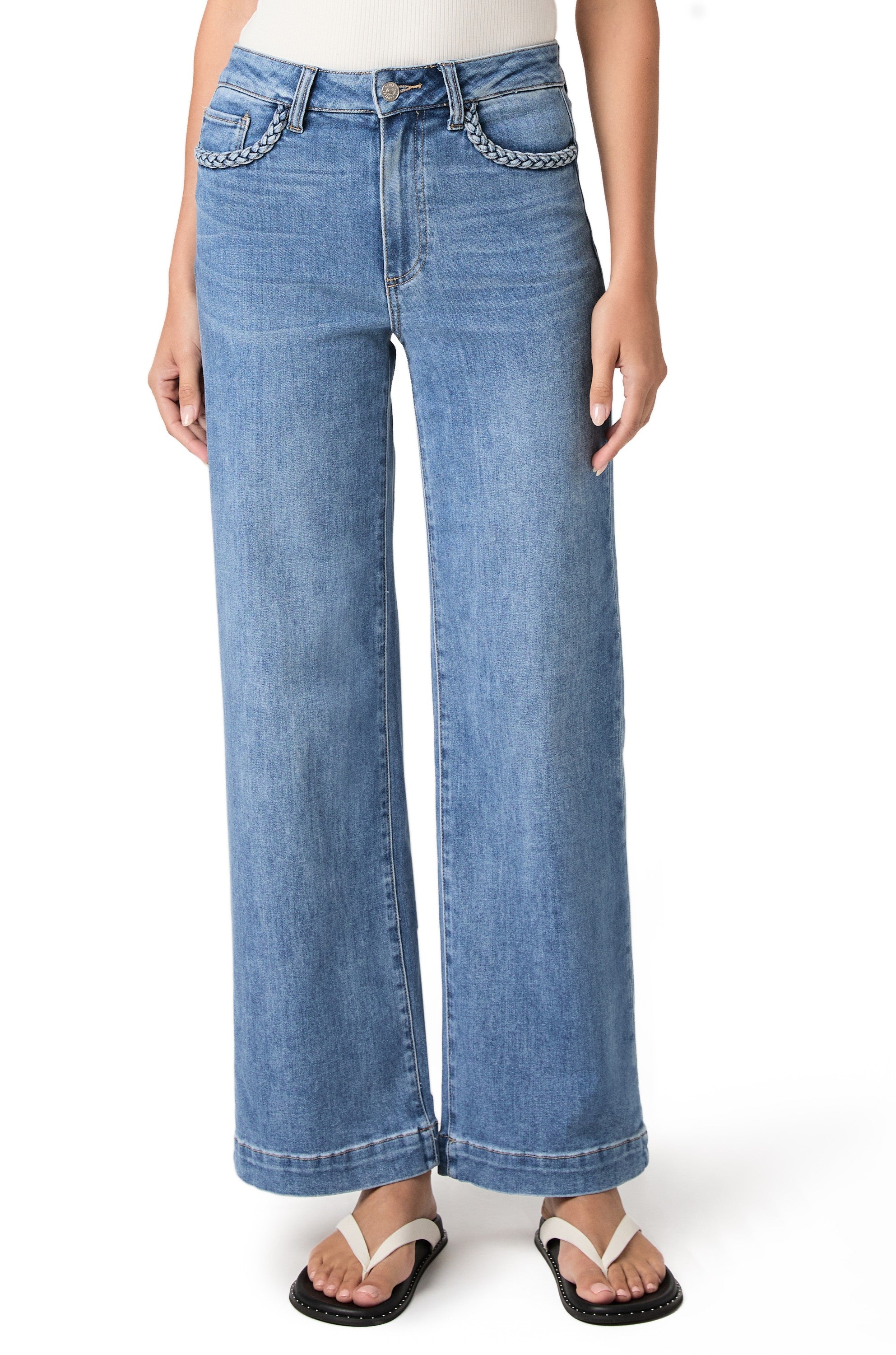 A woman stands against a white background wearing the Paige Anessa 31" w/ Braided Pocket Edge in Encino—light blue high-rise wide leg jeans featuring braided detailing on the front pockets and crafted from TRANSCEND VINTAGE denim—paired with a white ribbed tank top and white sandals.