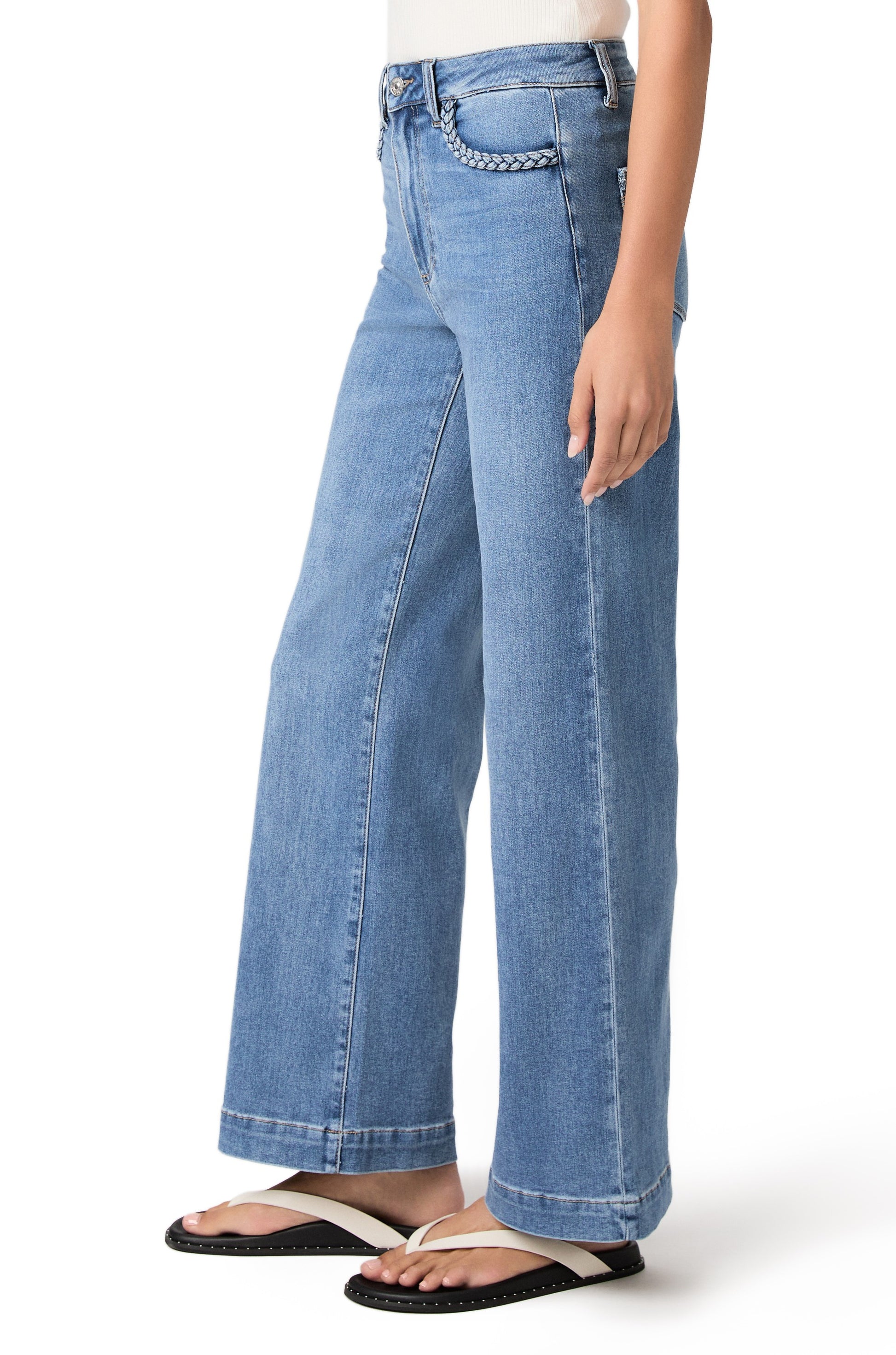 A person wearing PAIGE Anessa 31" with Braided Pocket Edge jeans in the Encino wash, a white sleeveless top, and black sandals stands against a white background. Only the lower half of the body is visible.