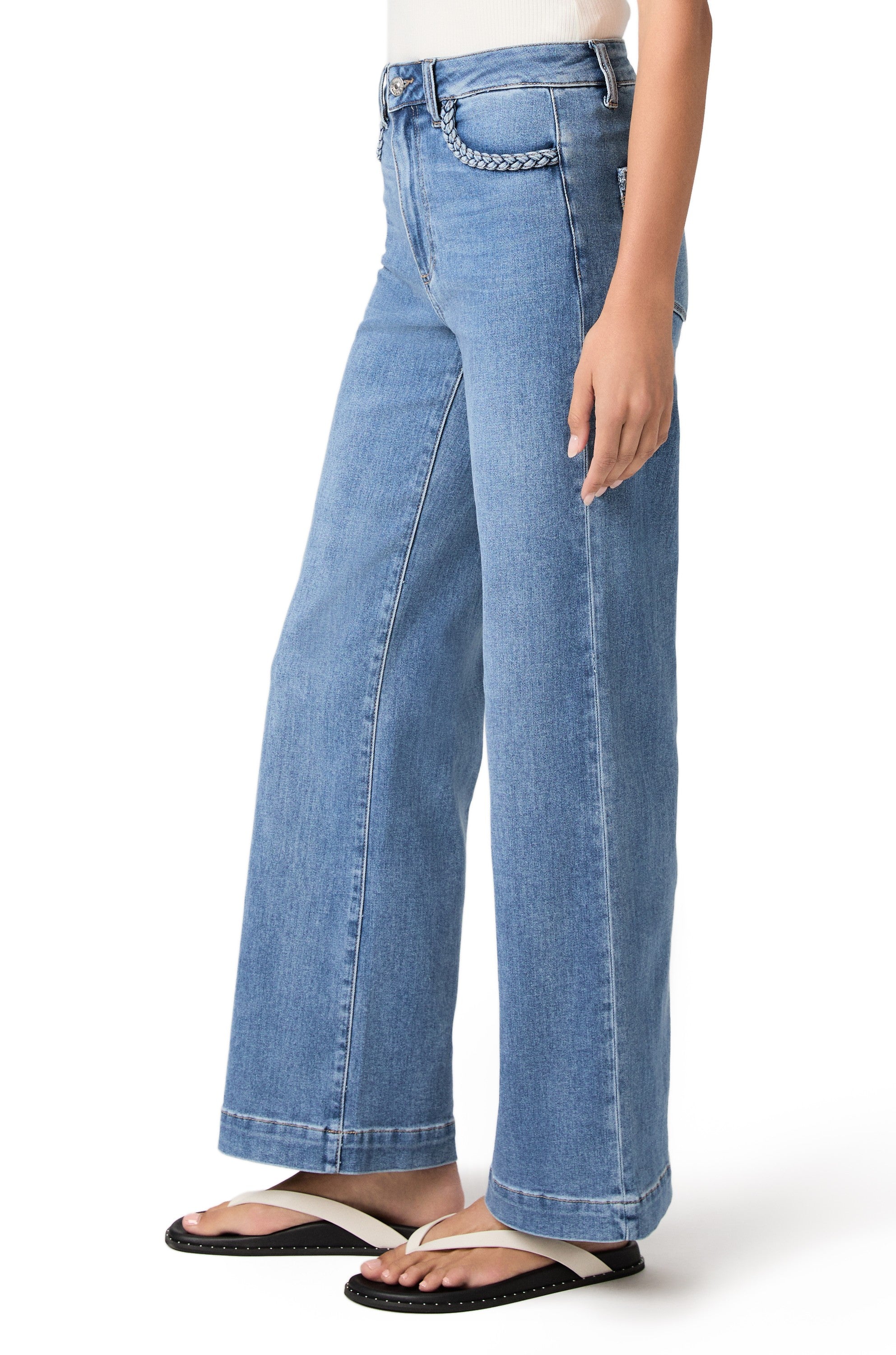 A person wearing PAIGE Anessa 31" with Braided Pocket Edge jeans in the Encino wash, a white sleeveless top, and black sandals stands against a white background. Only the lower half of the body is visible.