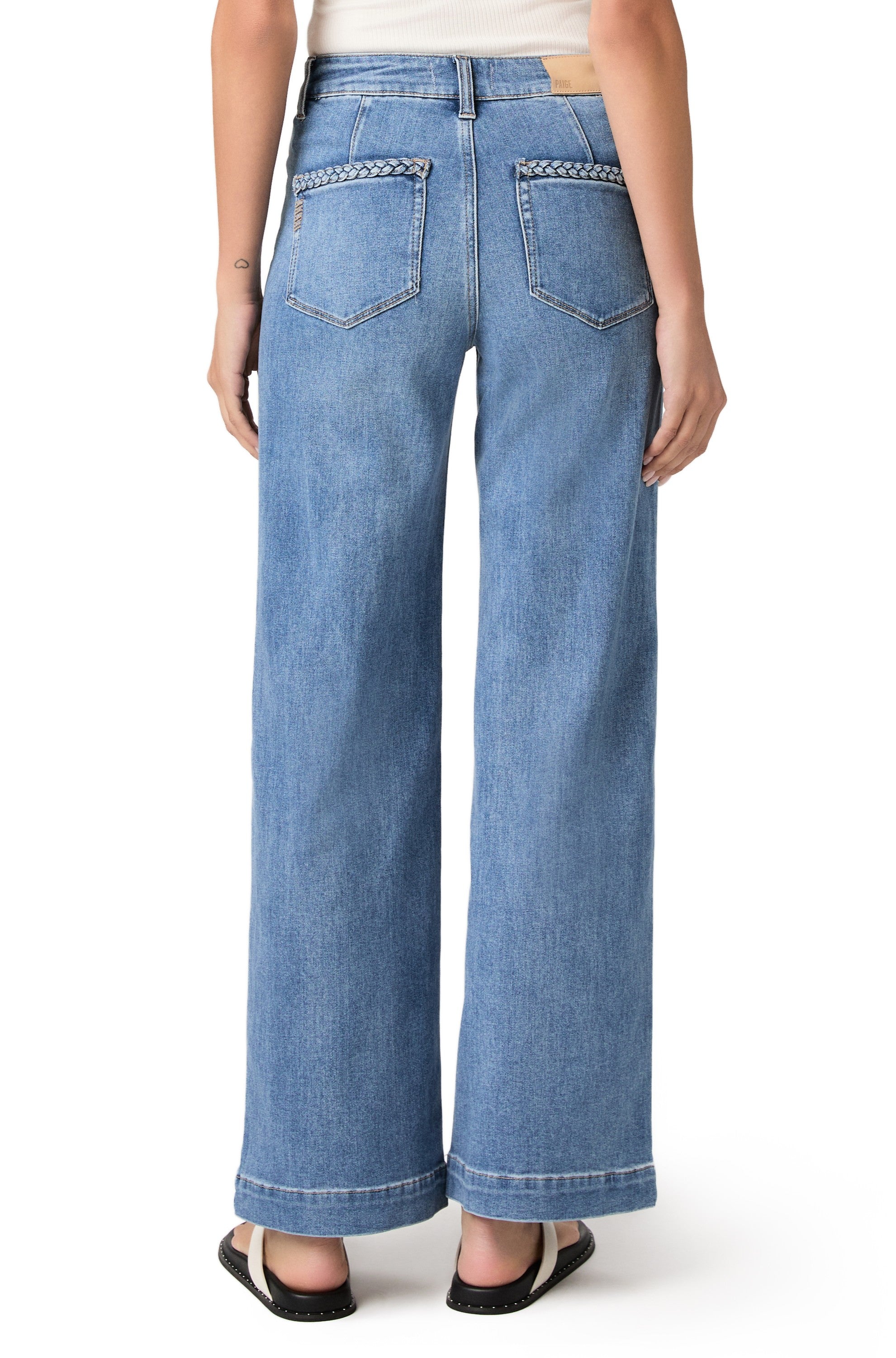 A person is shown from the back against a white background, wearing the Paige Anessa 31" w/ Braided Pocket Edge in Encino—high rise wide leg jeans made from TRANSCEND VINTAGE denim with braided detailing on the back pockets—paired with a white top and black sandals.