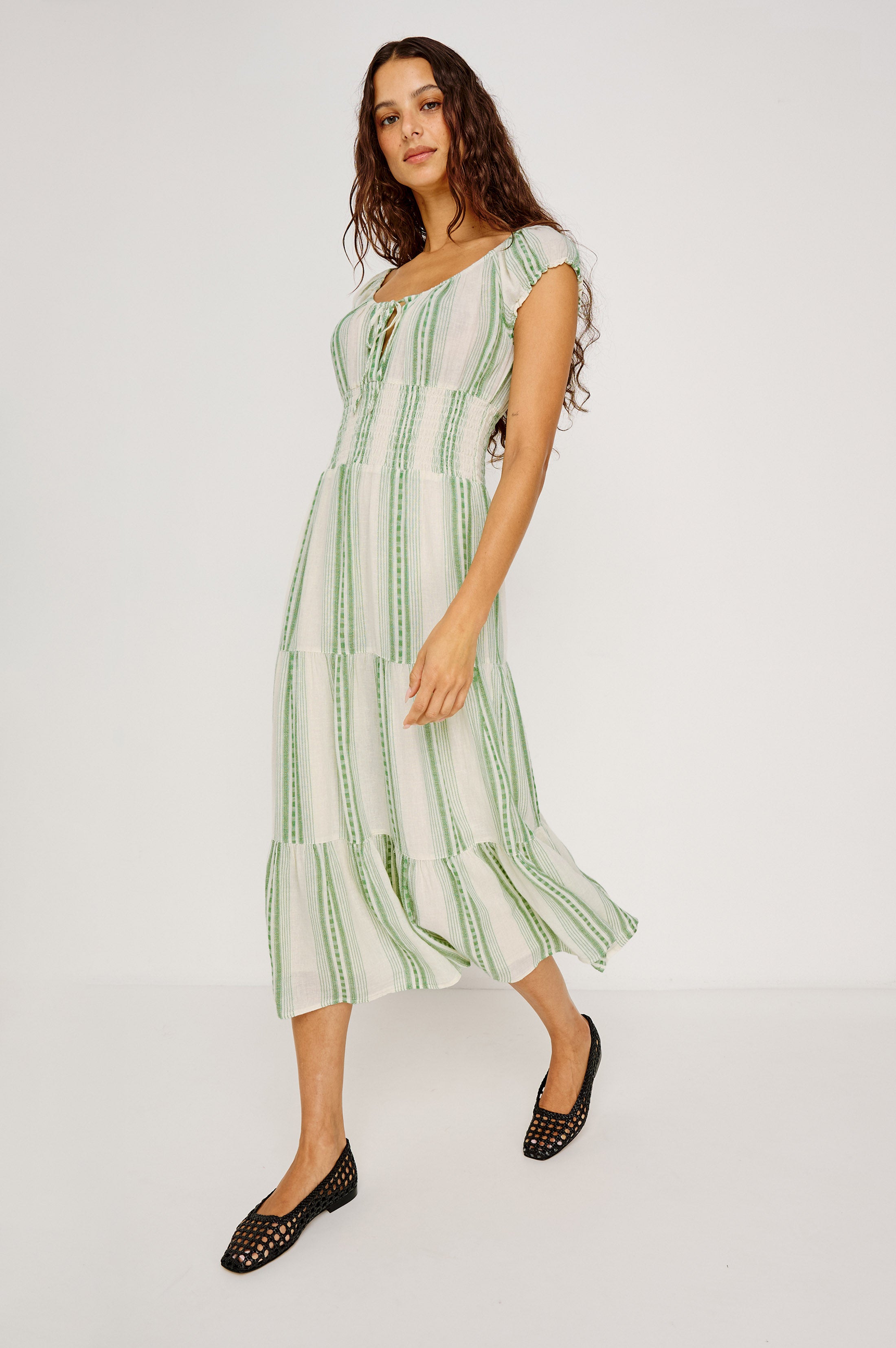 A woman with long, wavy hair wears the Rails Daria Midi Dress—a light, short-sleeve linen dress with green stripes and a tiered skirt—paired with black woven flats as she walks against a plain white background.