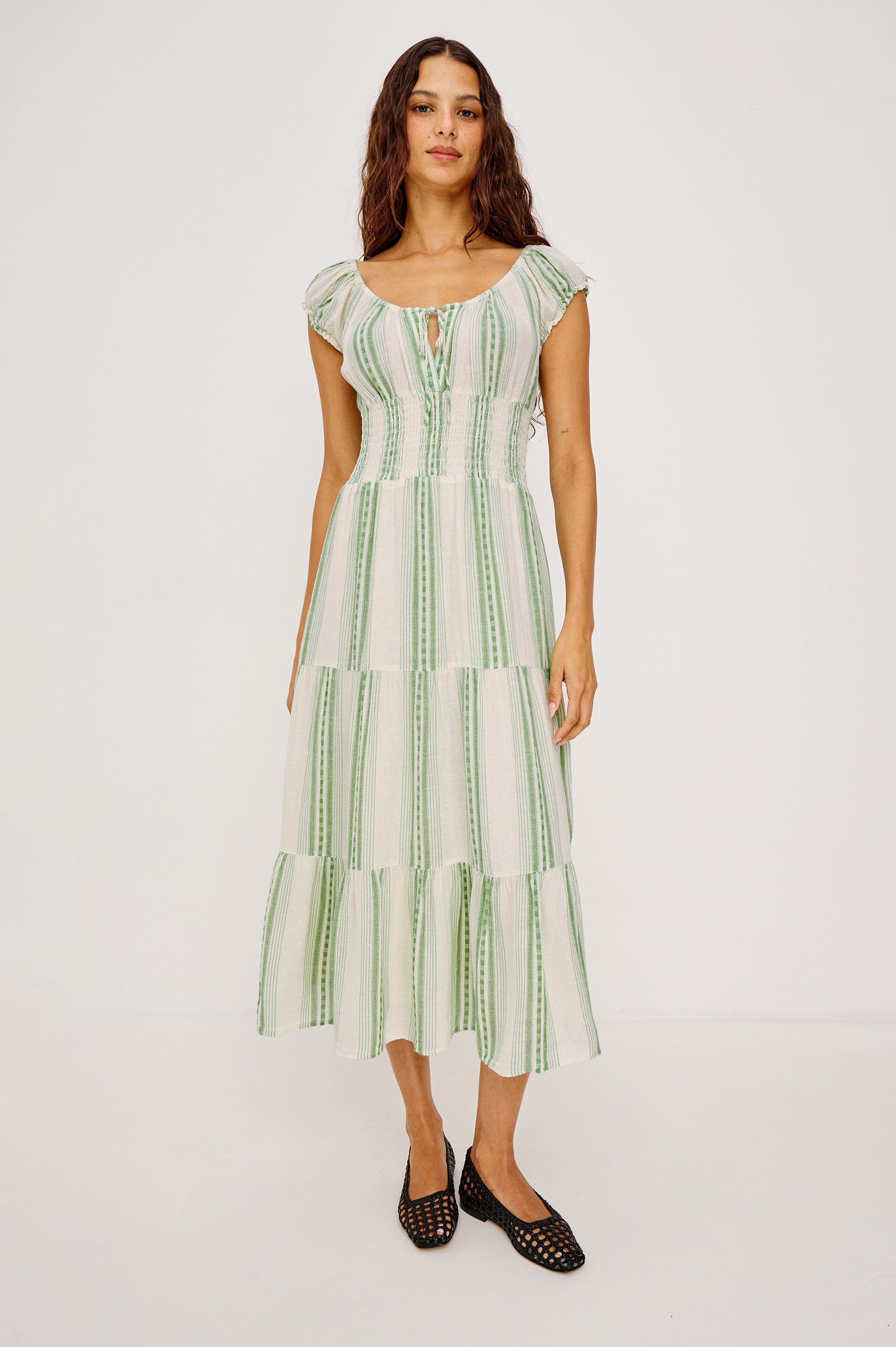 A woman with loose, wavy hair and a neutral expression wears the Rails Daria Midi Dress in Green Multi, featuring green vertical stripes, short sleeves, and a gathered bust, paired with black woven shoes against a plain backdrop.