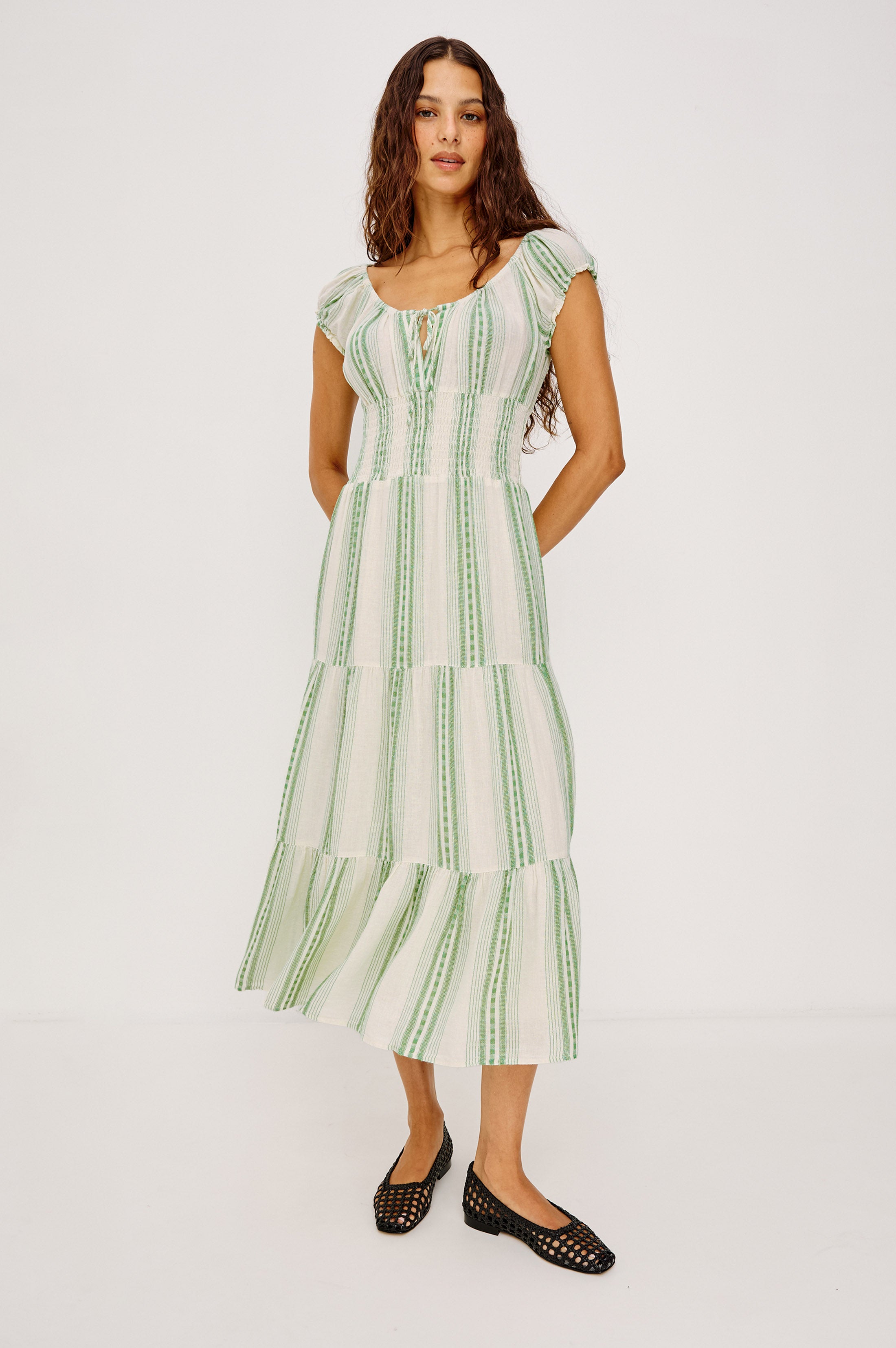 A woman with long, wavy brown hair and a neutral expression wears the Rails Daria Midi Dress—lightweight with green vertical stripes, flutter sleeves, and a tiered skirt—paired with black woven flats against a plain white background.