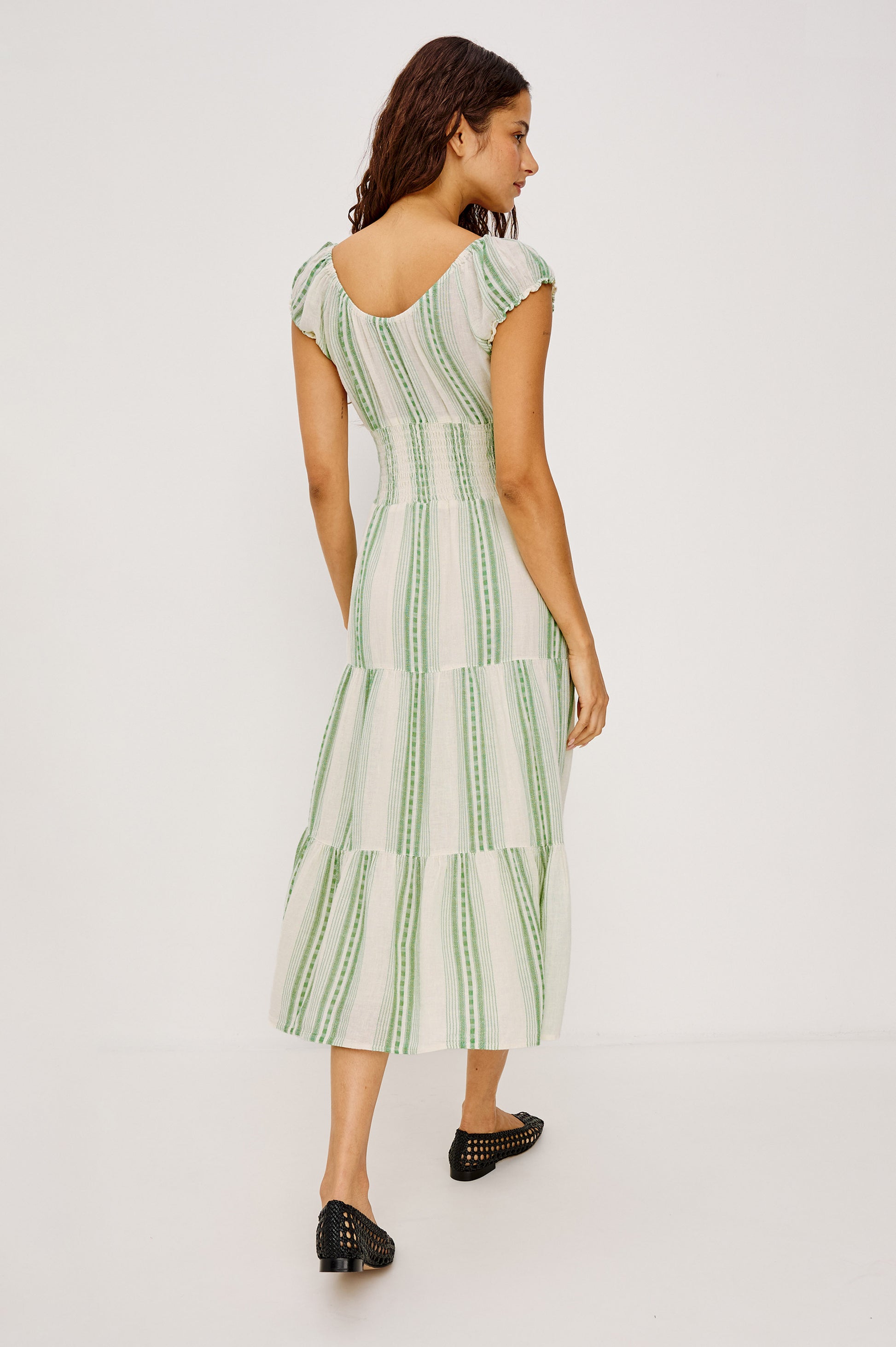 A woman with wavy brown hair wears the Rails Daria Midi Dress, a lightweight ankle-length linen dress with green vertical stripes and cap sleeves, paired with black flats. She faces away, looking left against a plain background.