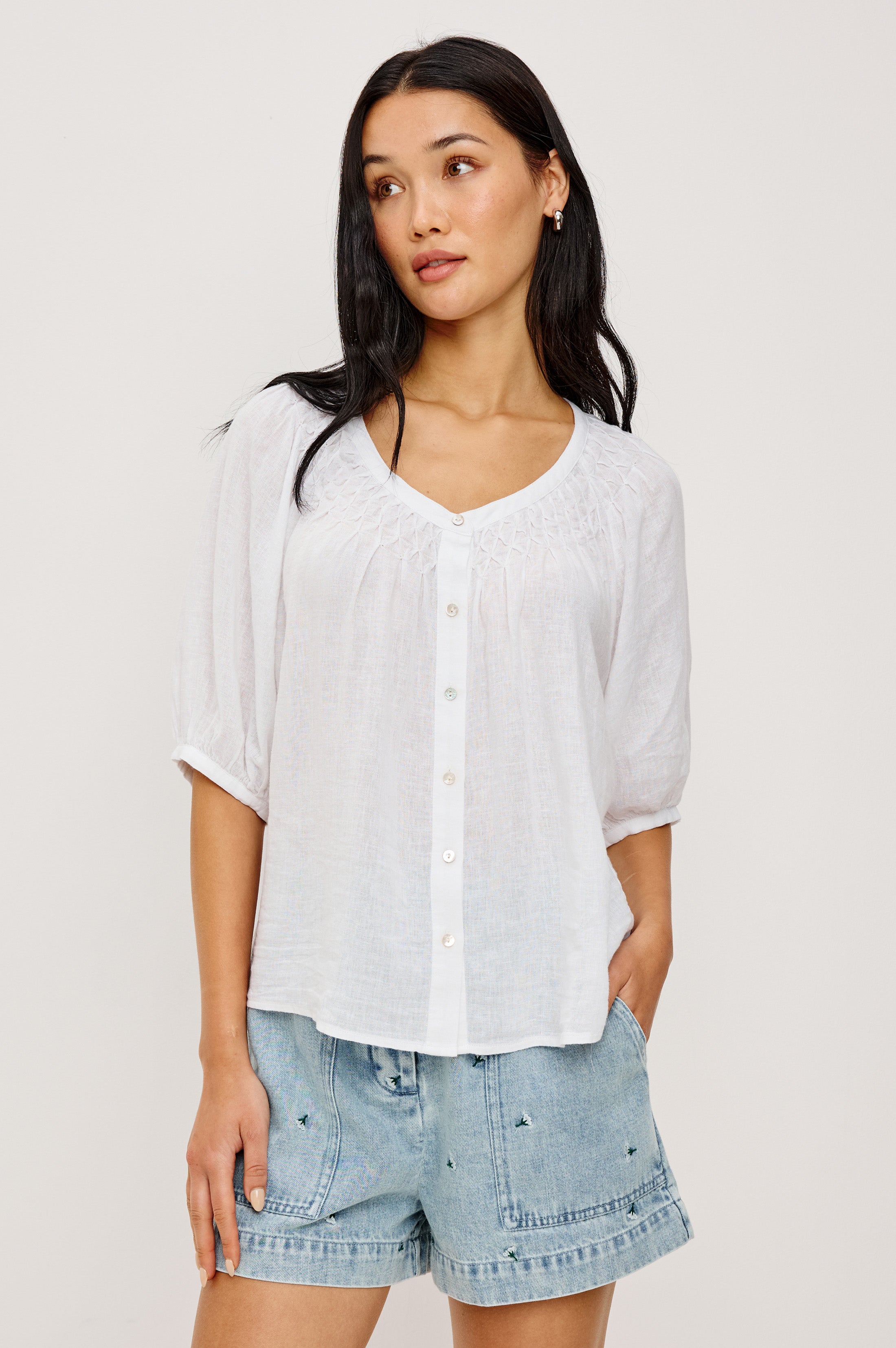 A woman with long dark hair wears the Rails Rix Blouse—a relaxed button-up with three-quarter sleeves—paired with light blue denim shorts featuring small embroidered details, standing against a plain light background.