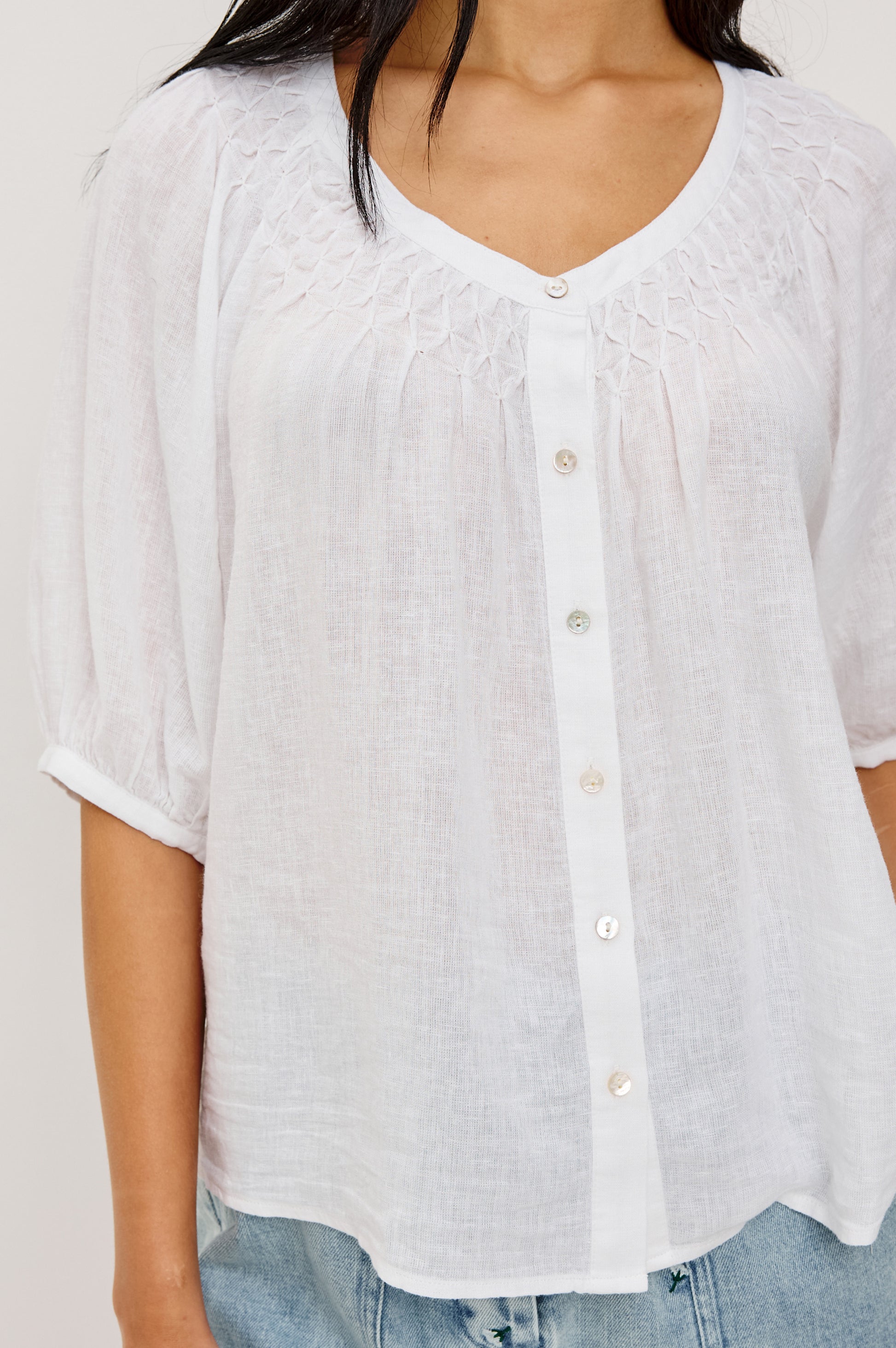 A woman wearing the Rails Rix Blouse—a loose-fitting, white luxe linen button-up with a smocked yoke and rolled sleeves—paired with light blue jeans. Photo is cropped below the shoulders.