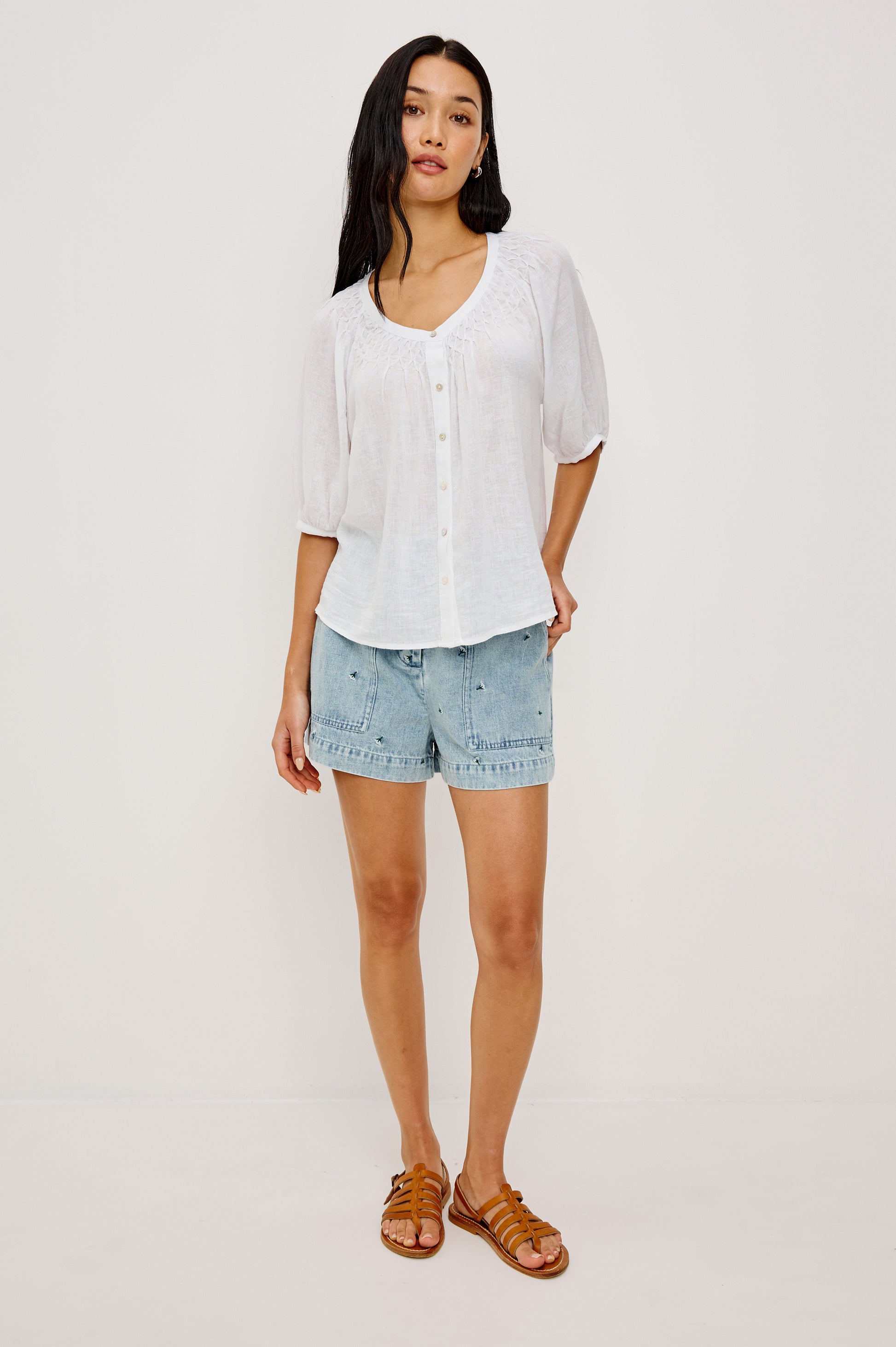 A woman with long dark hair poses casually against a white background, wearing the Rails Rix Blouse with elbow-length sleeves, paired with light denim shorts and brown flat sandals.