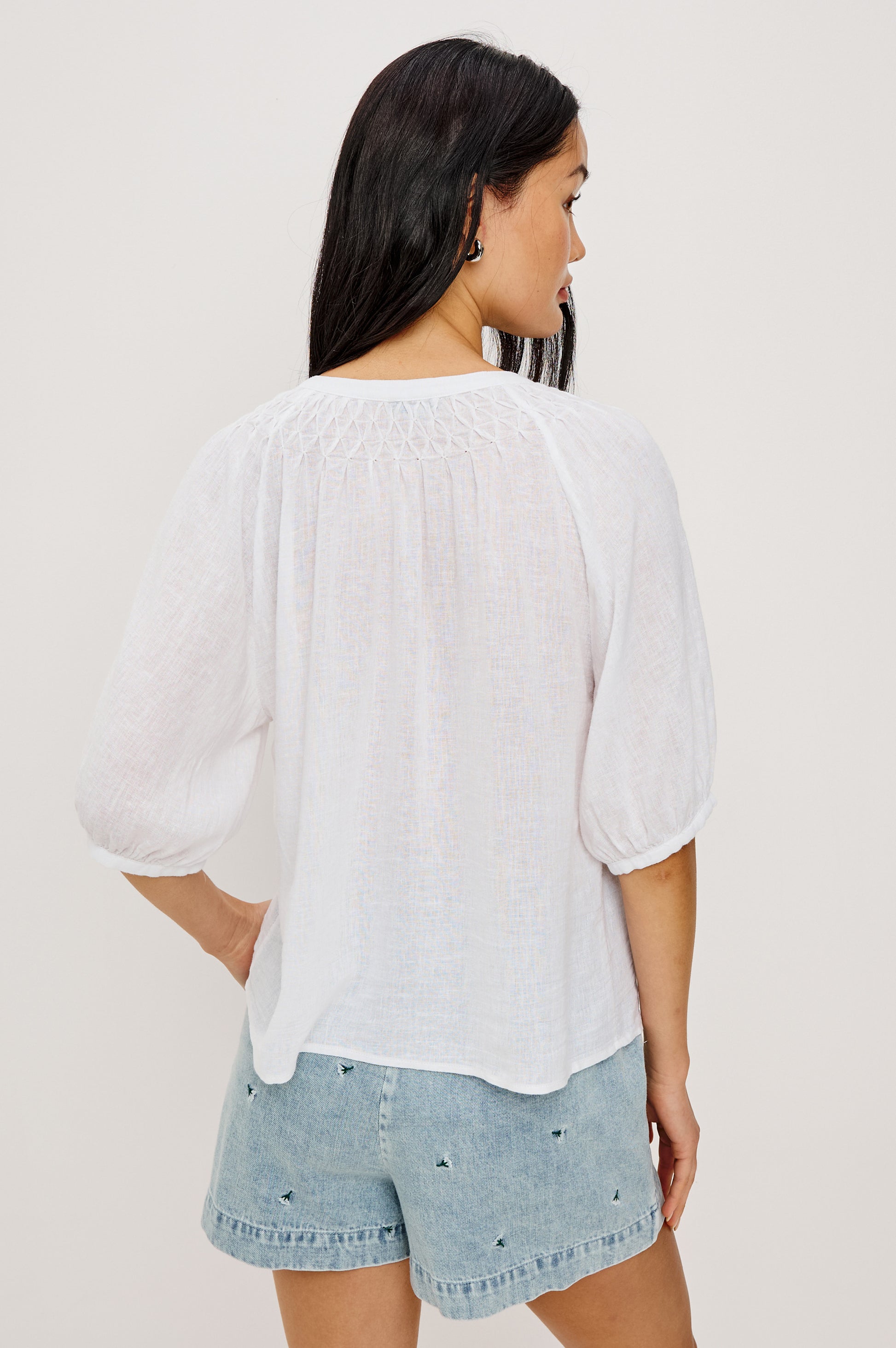 A woman with long dark hair faces away, wearing the Rails Rix Blouse—a relaxed button-up with three-quarter sleeves—paired with light blue denim shorts featuring small embroidered details. Her left hand is on her hip against a plain background.
