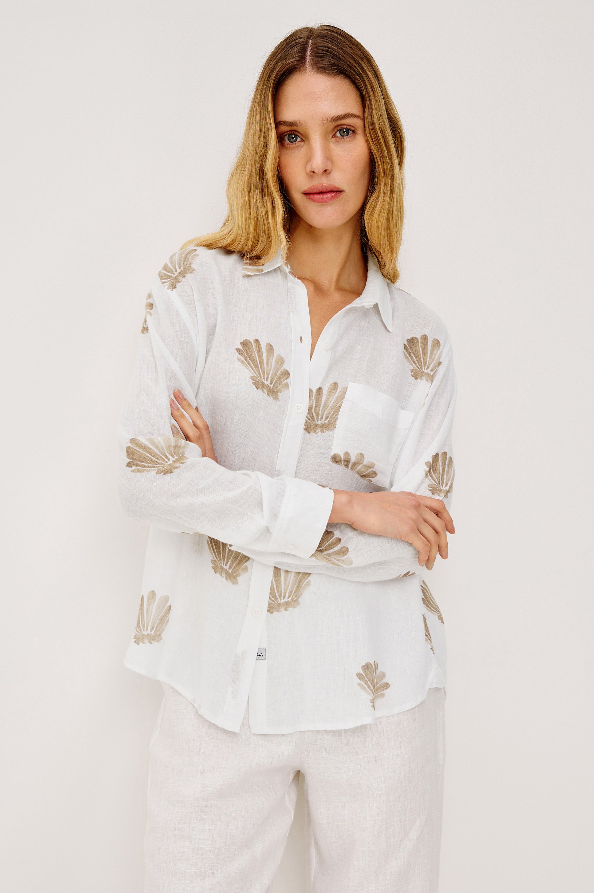 A woman with long, wavy blonde hair models the Rails Charli Shell Top—a relaxed-fit white linen shirt with a beige seashell print—paired with white pants, standing against a light, plain background.