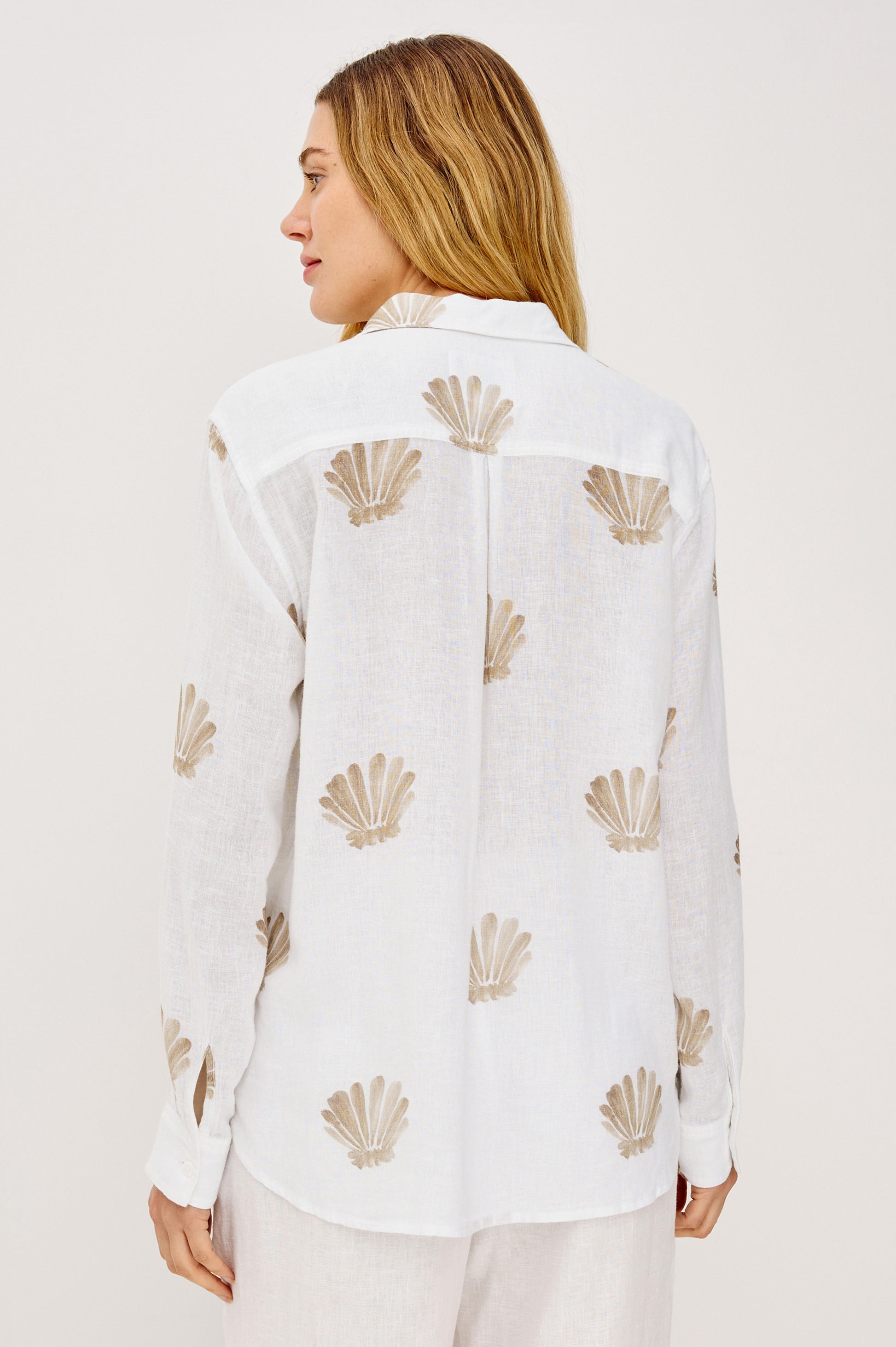 A woman with long, light brown hair is seen from behind wearing the Rails Charli Shell Top—a relaxed fit white linen shirt with a beige seashell pattern—perfect for layering and standing out against a plain, light background.