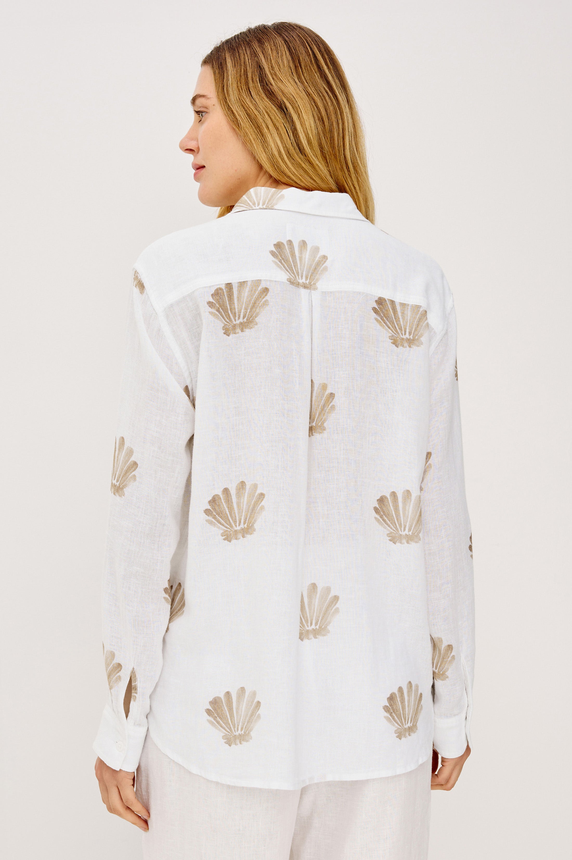 A woman with long, light brown hair is seen from behind wearing the Rails Charli Shell Top—a relaxed fit white linen shirt with a beige seashell pattern—perfect for layering and standing out against a plain, light background.