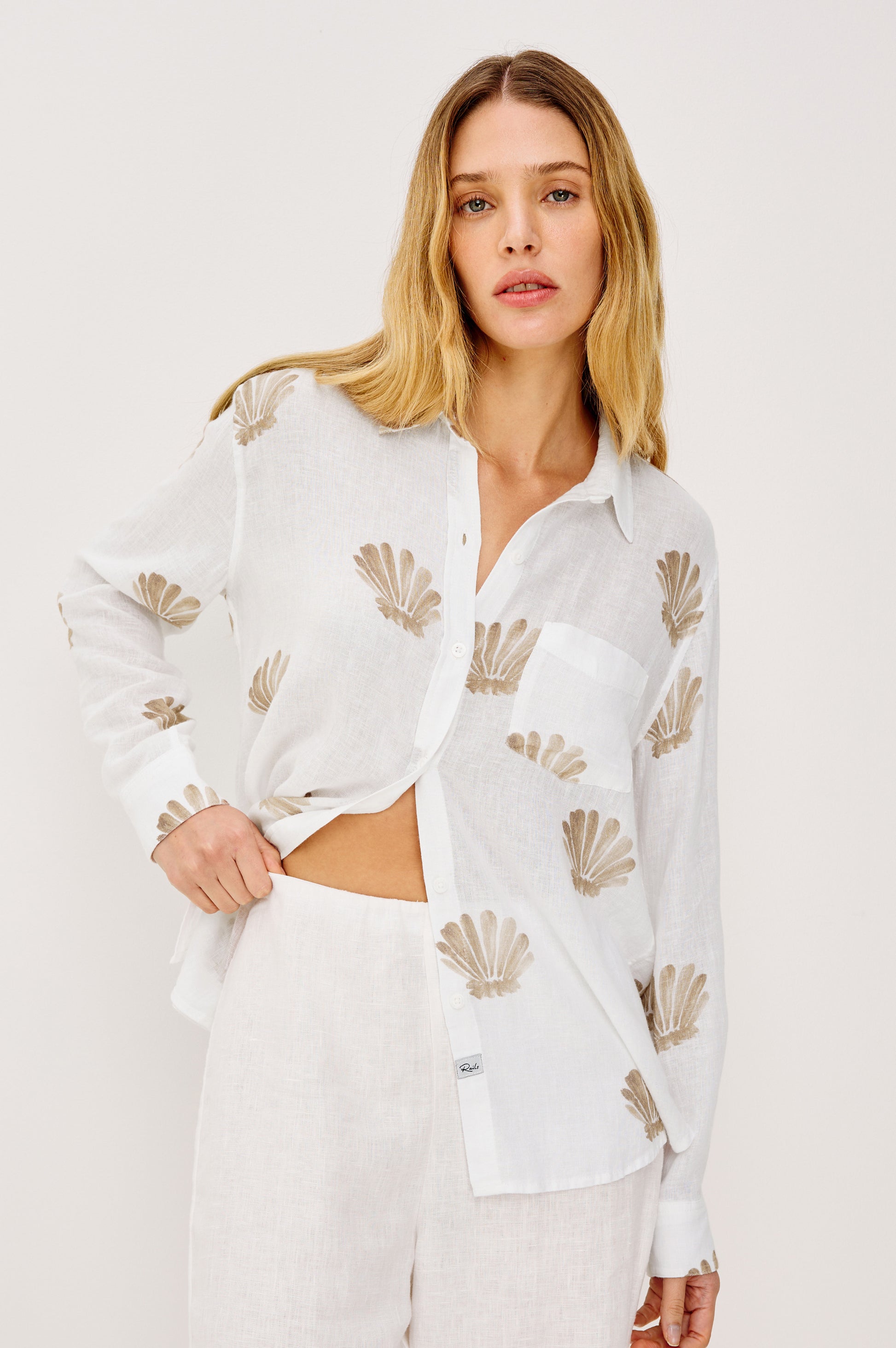 A woman with long blonde hair wears the Rails Charli Shell Top, a relaxed fit linen shirt with beige shell patterns, paired with white pants, standing against a plain light background with a relaxed expression.