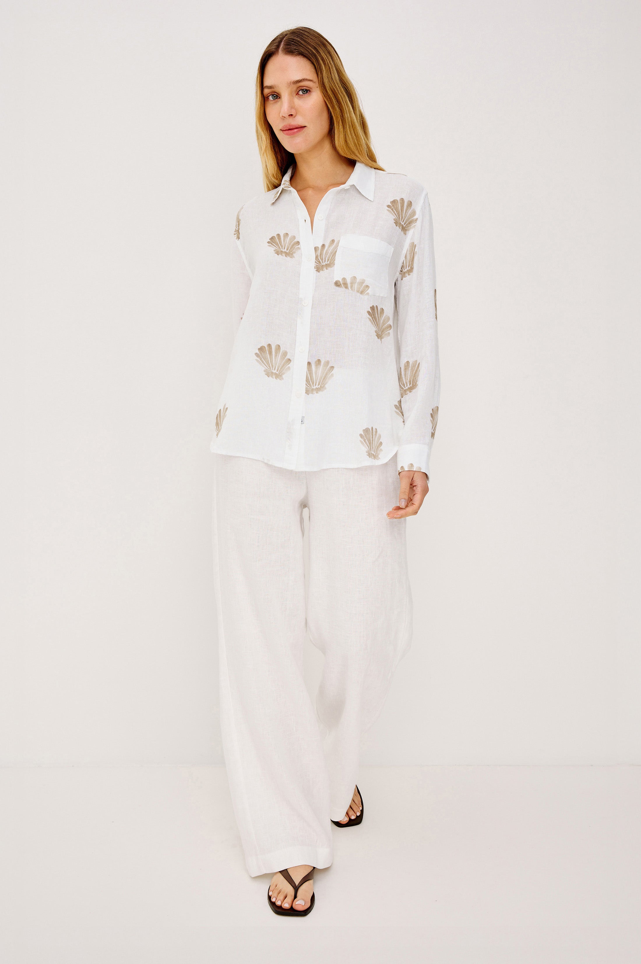 A woman models the Rails Charli Shell Top, a white long-sleeve linen shirt with beige shell patterns, paired with loose white pants and black sandals. The relaxed fit is ideal for effortless layering against a plain white background.